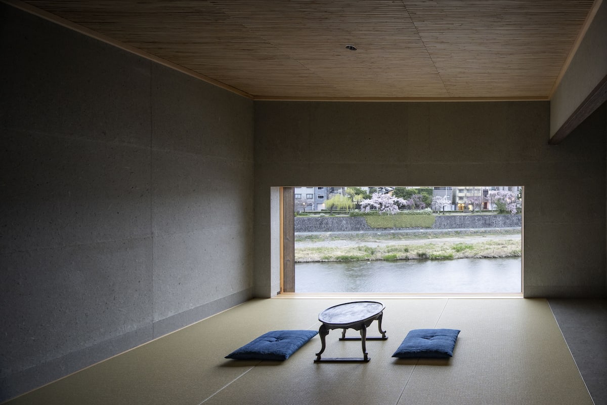 A room with tatami flooring, a low table, and cushions. A large window offers a view of a water body and greenery.