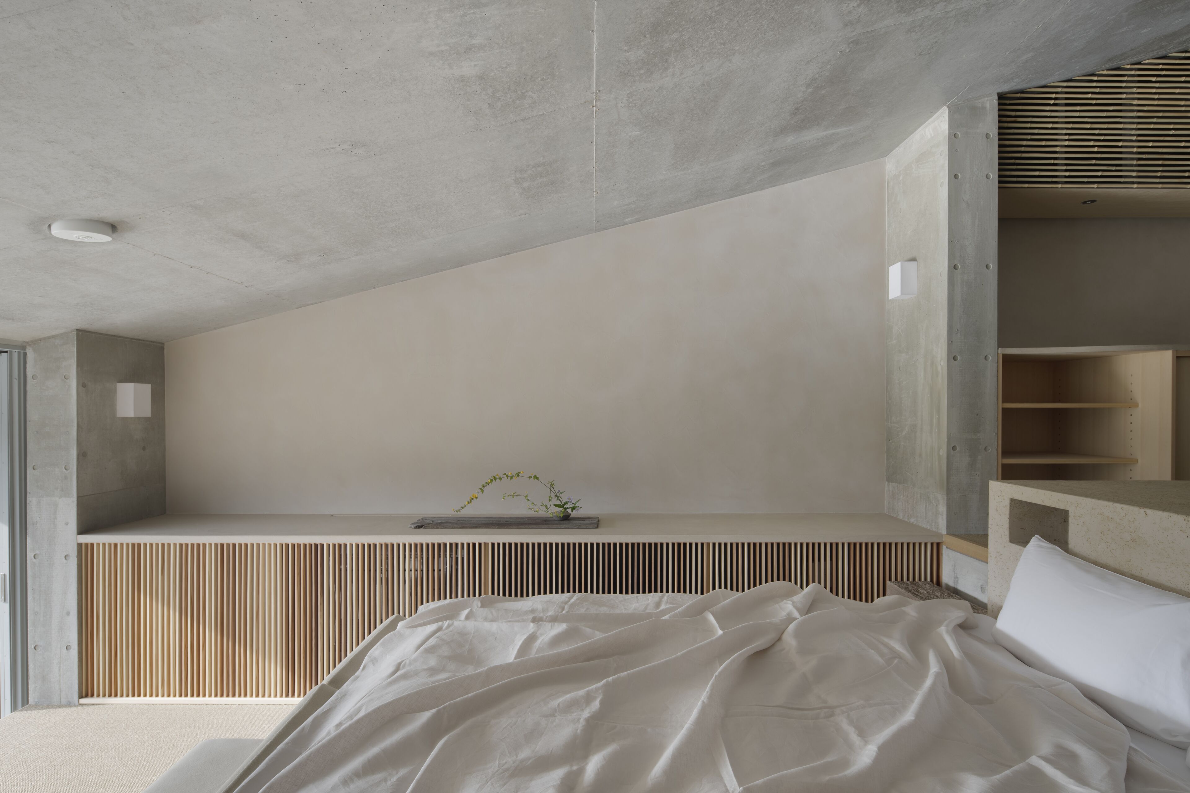 A modern bedroom with a concrete ceiling, a slatted wooden bench, and a bed with white bedding.