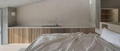 A modern bedroom with a concrete ceiling, a slatted wooden bench, and a bed with white bedding.