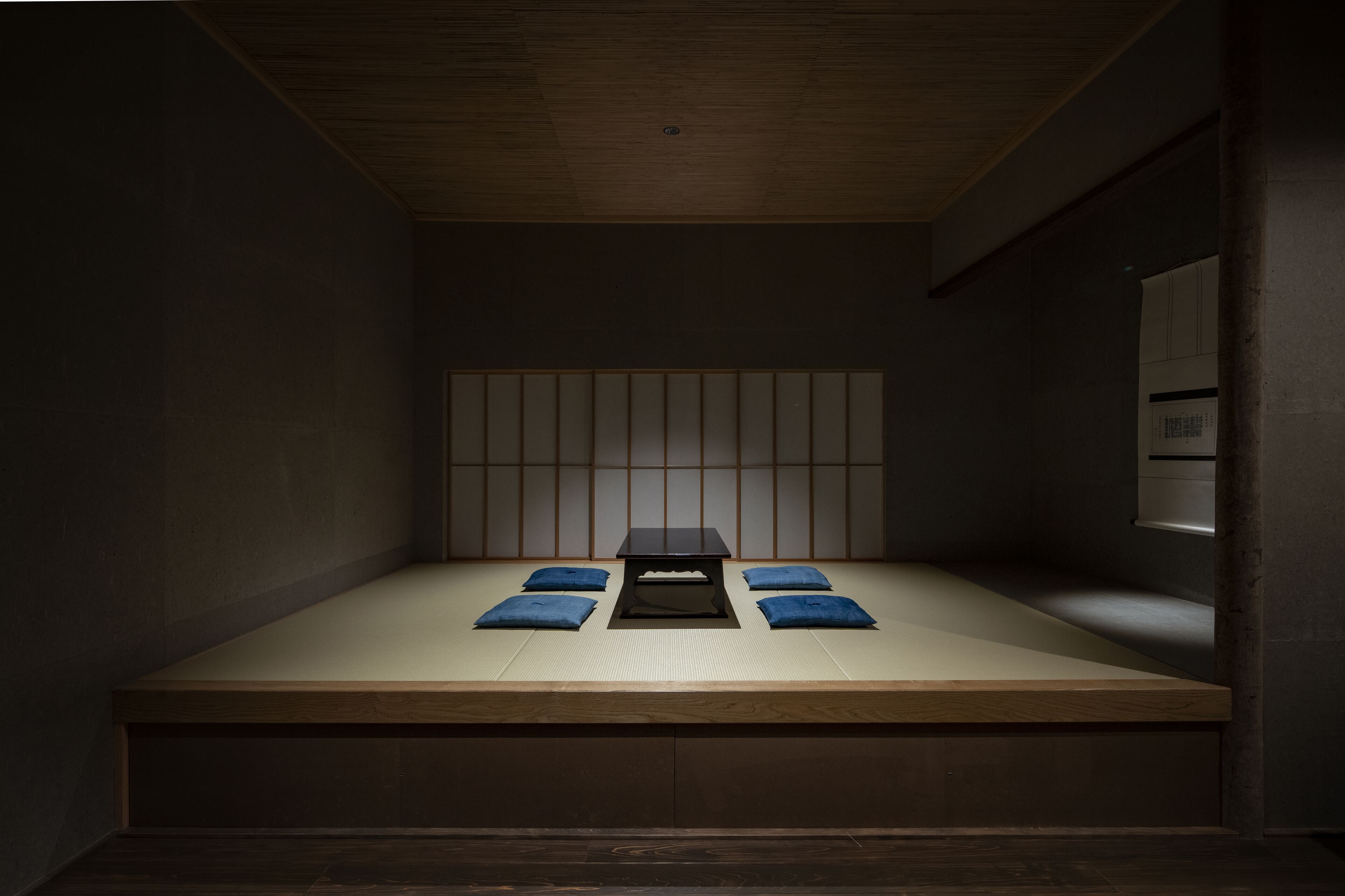 Habitació amb un llit de fusta baix, un paviment de tatami, una paret amb panells shoji i una taula petita amb coixins.