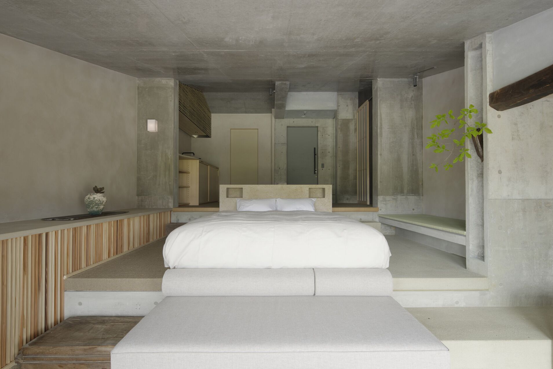 Un dormitorio moderno con una cama grande, una mesita de noche pequeña, un banco y vistas a un pasillo.