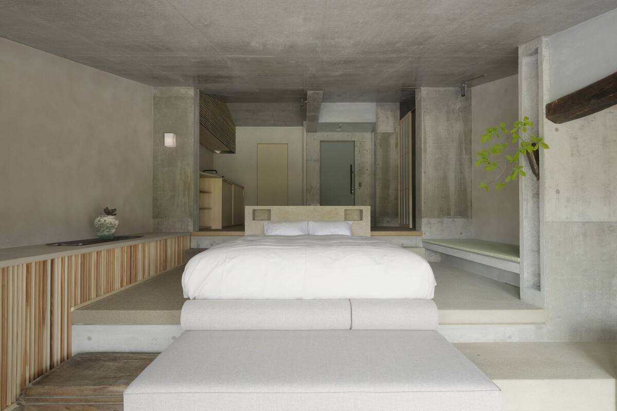 Un dormitorio moderno con una cama grande, una mesita de noche pequeña, un banco y vistas a un pasillo.
