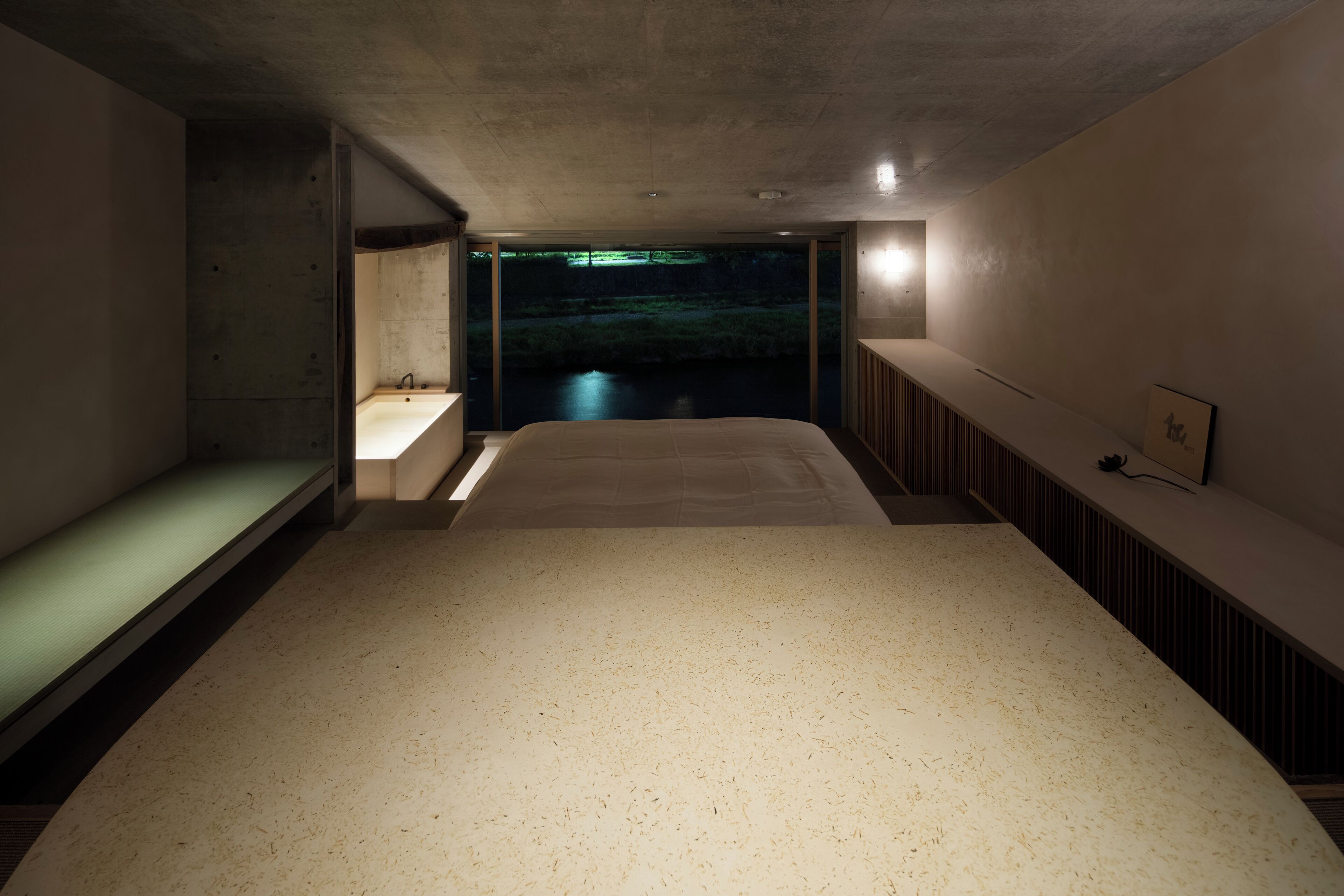 A modern, minimalist bedroom with a large bed, a built-in sink, and a view of a water body.