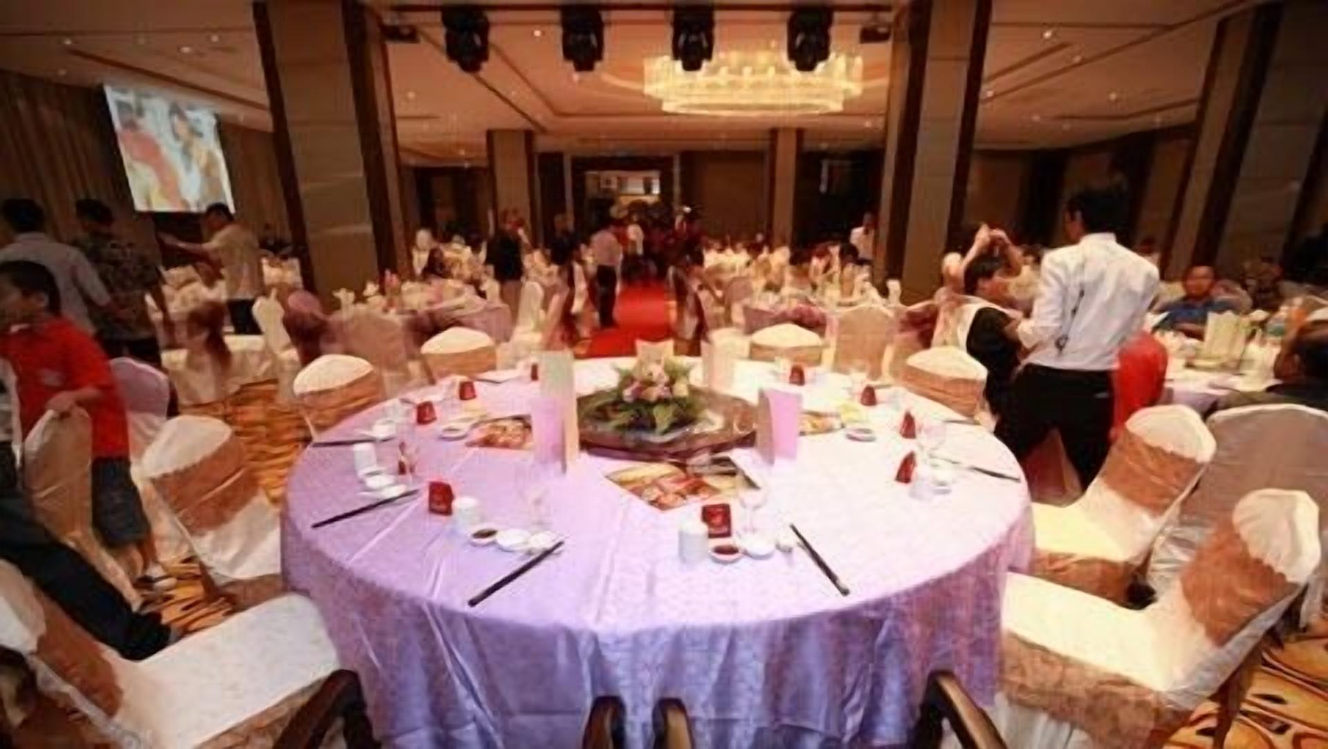Une salle de banquet avec des tables rondes recouvertes de nappes blanches, entourées de chaises blanches ornées de rubans rouges.
