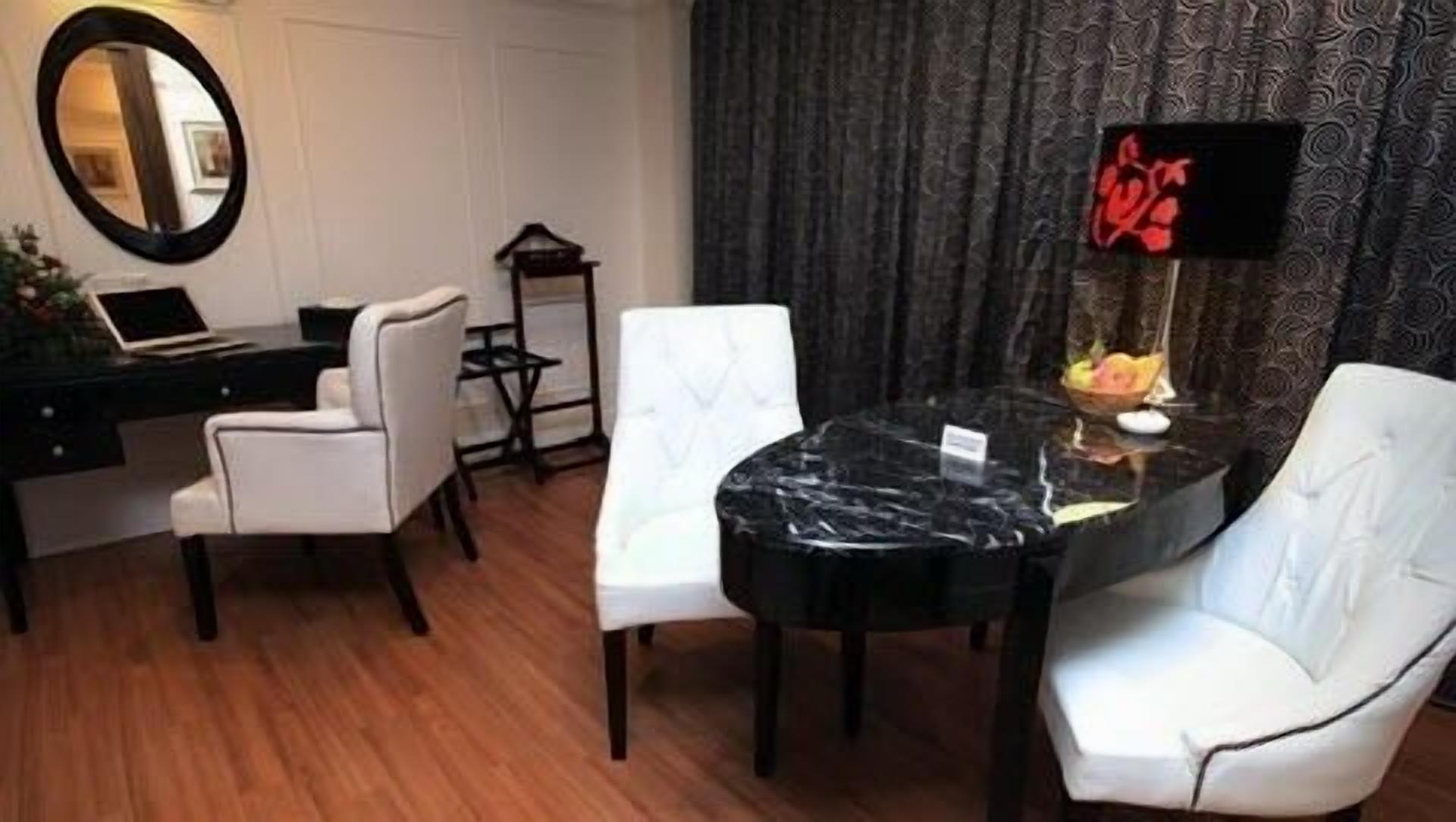 A hotel room with a dining area featuring a marble table, white upholstered chairs, a laptop on a desk, and a decorative mirror.