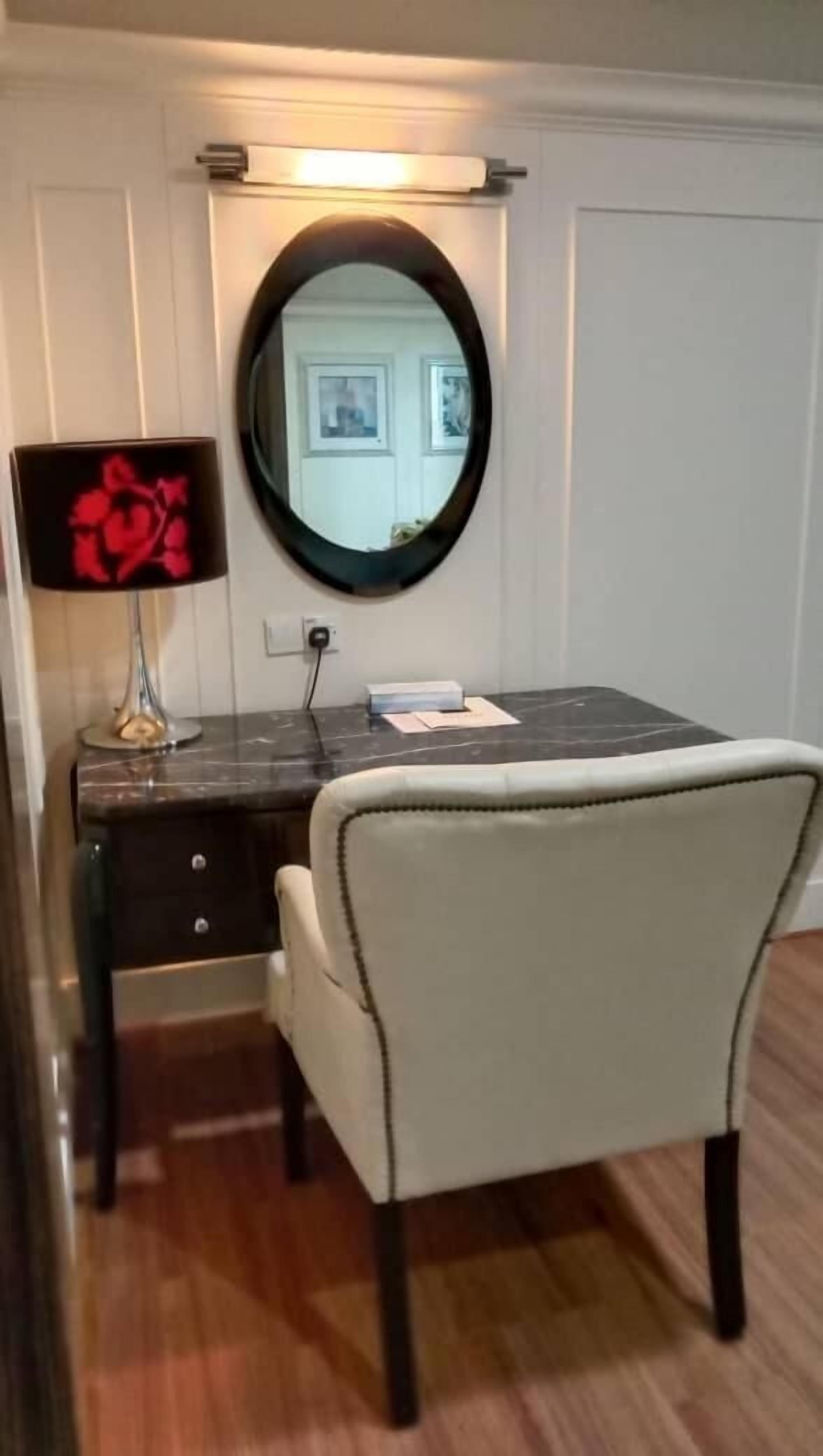 Una habitación con un escritorio de mármol oscuro, una silla blanca, un espejo redondo y una lámpara con un diseño floral rojo.