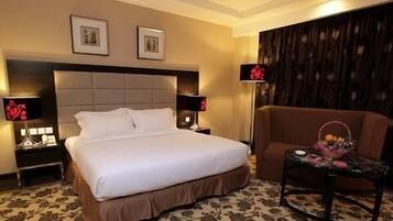 Una habitación de hotel con una cama grande, dos mesitas de noche con lámparas, un sillón marrón y una pequeña mesa con una cesta.