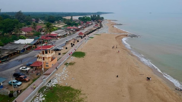 A beach with a road running parallel to it, lined with buildings and parked cars.