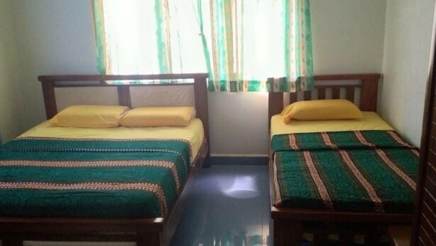 Una habitación con dos camas individuales, cada una con cabecero de madera y colcha estampada en verde y naranja. Hay una ventana con cortinas verdes y blancas, y el suelo es de baldosas.