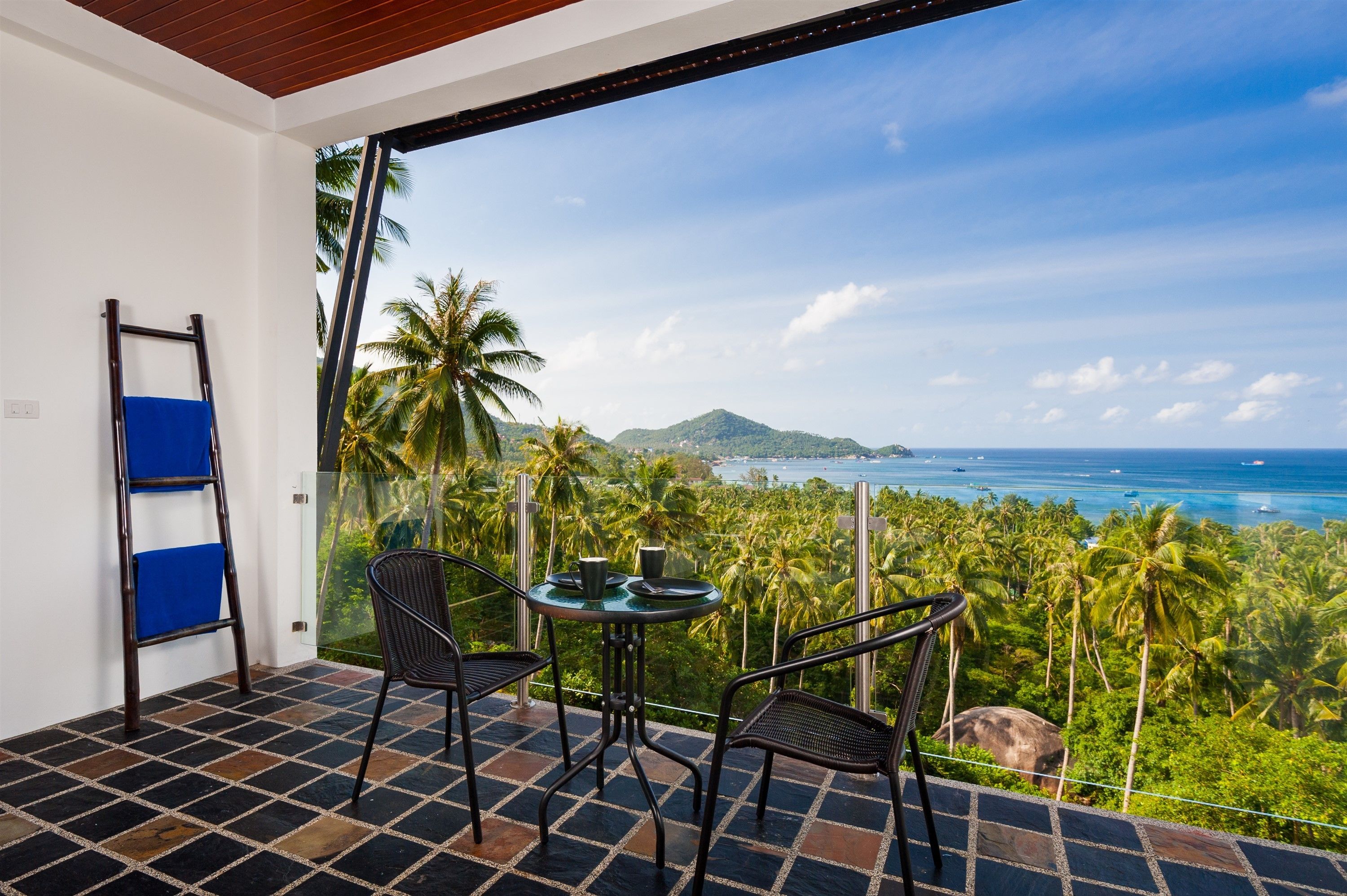 A balcony with a table and chairs, overlooking a coastal landscape with palm trees and a view of the sea.