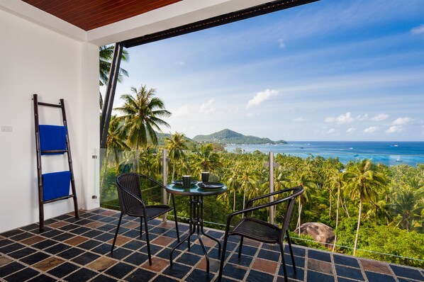 Un balcon avec une table et des chaises, donnant sur un paysage côtier avec des palmiers et une vue sur la mer.