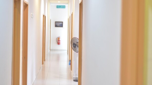 Un pasillo largo y estrecho con puertas de madera a ambos lados, un extintor en la pared y un ventilador de techo a la derecha.