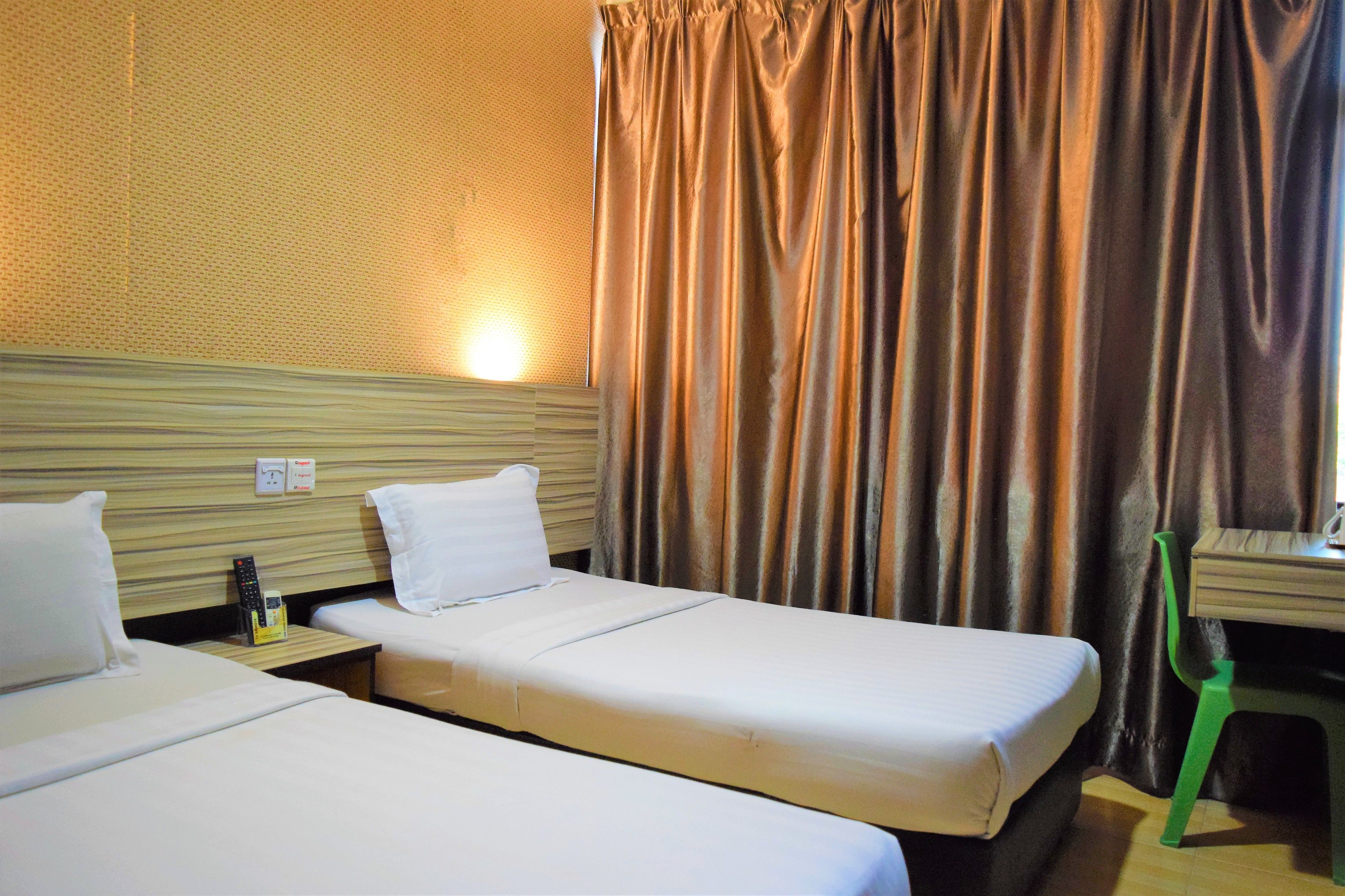 Habitación de hotel con dos camas individuales, un cabecero de madera, una mesita de noche con un control remoto, una silla verde y una cortina.