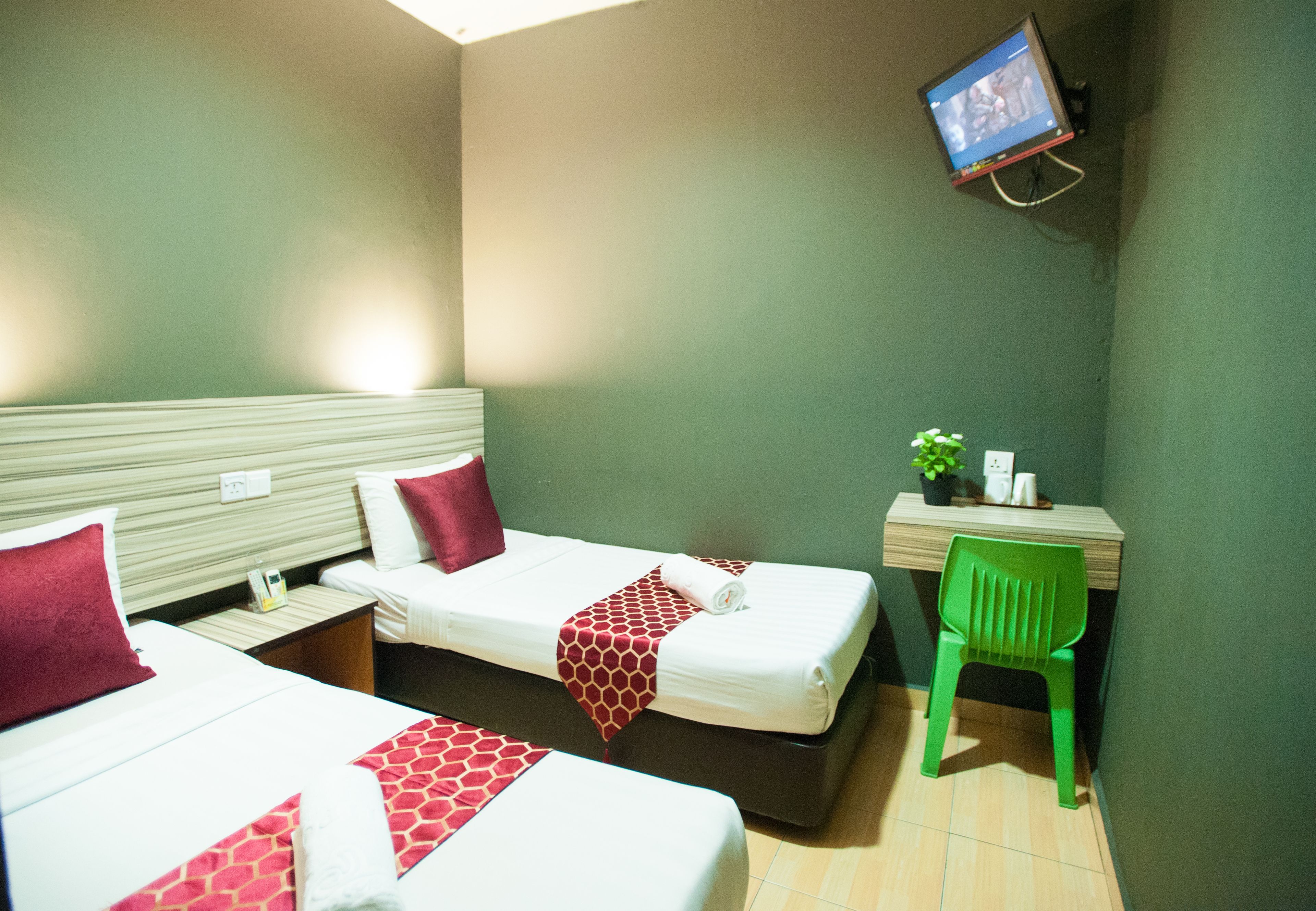 A hotel room with two beds, a desk, a green chair, and a television on the wall.