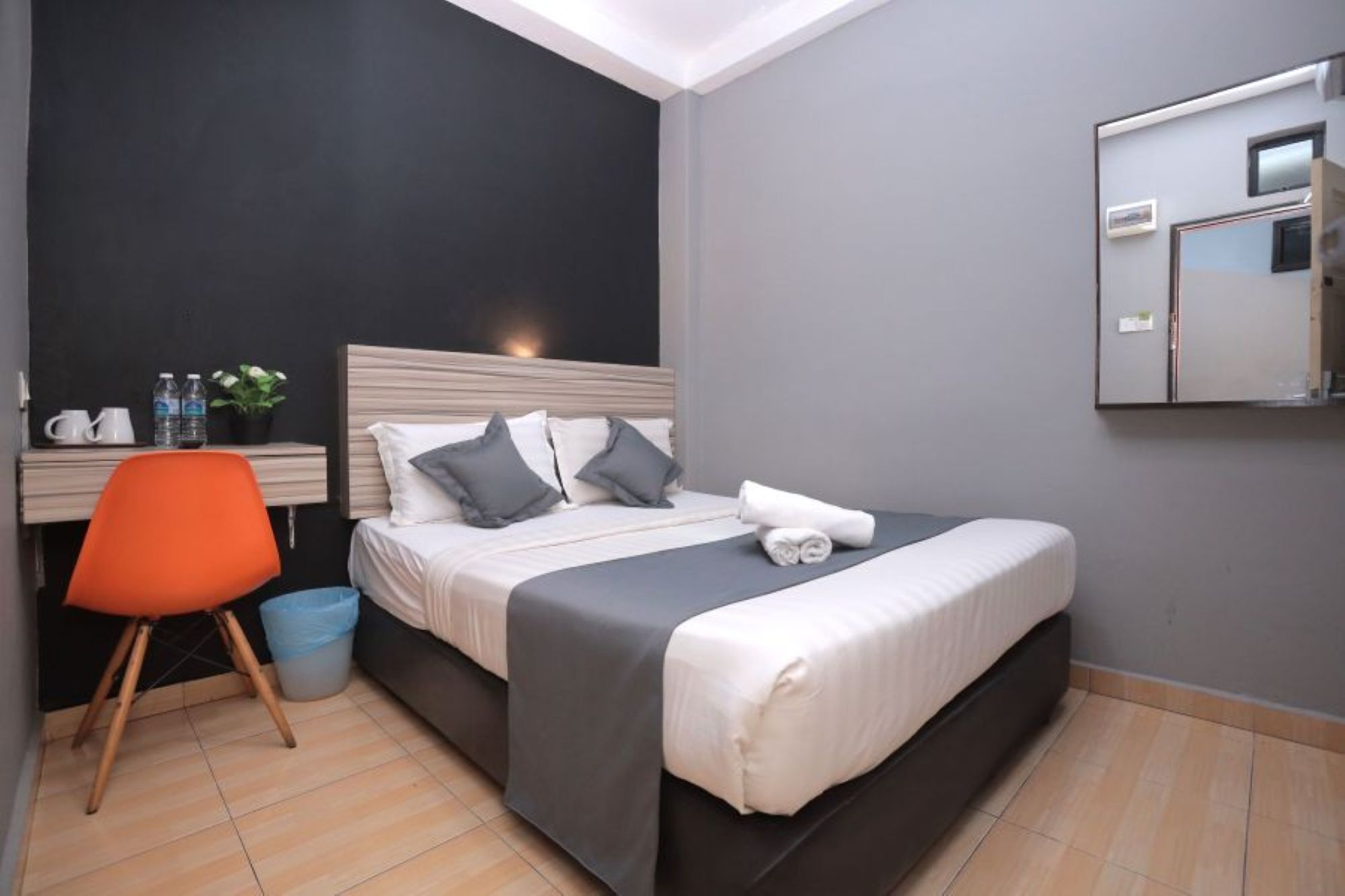 A hotel room with a bed, a desk with a chair, a mirror, and a small trash bin.