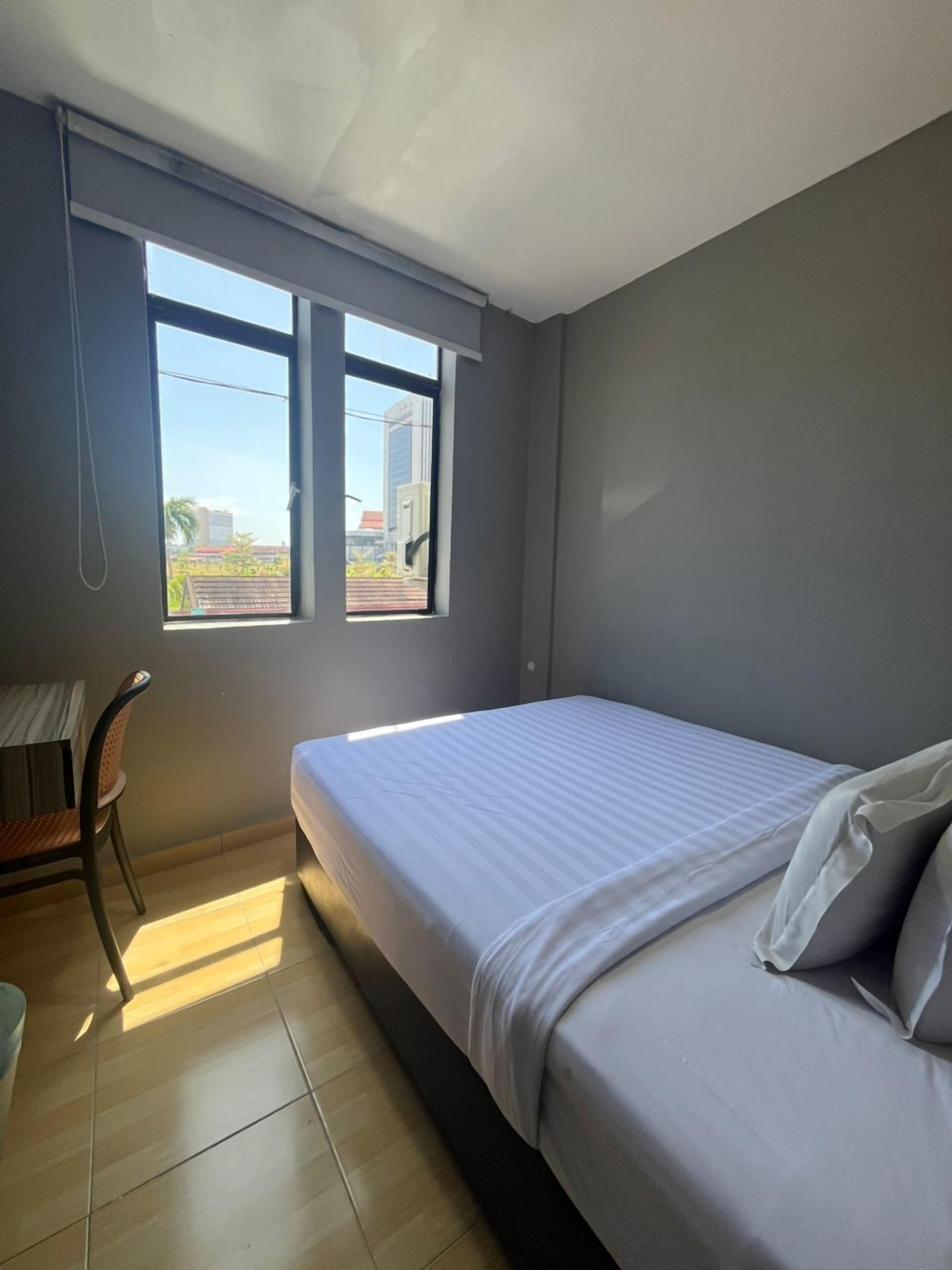 Ein Schlafzimmer mit einem Bett, einem Schreibtisch und einem Stuhl. Zwei Fenster sorgen für natürliches Licht.