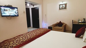 A bedroom with a large bed, a television mounted on the wall, a chair, and a desk with a lamp. - Hotel Shubham Majesty (Mathura)