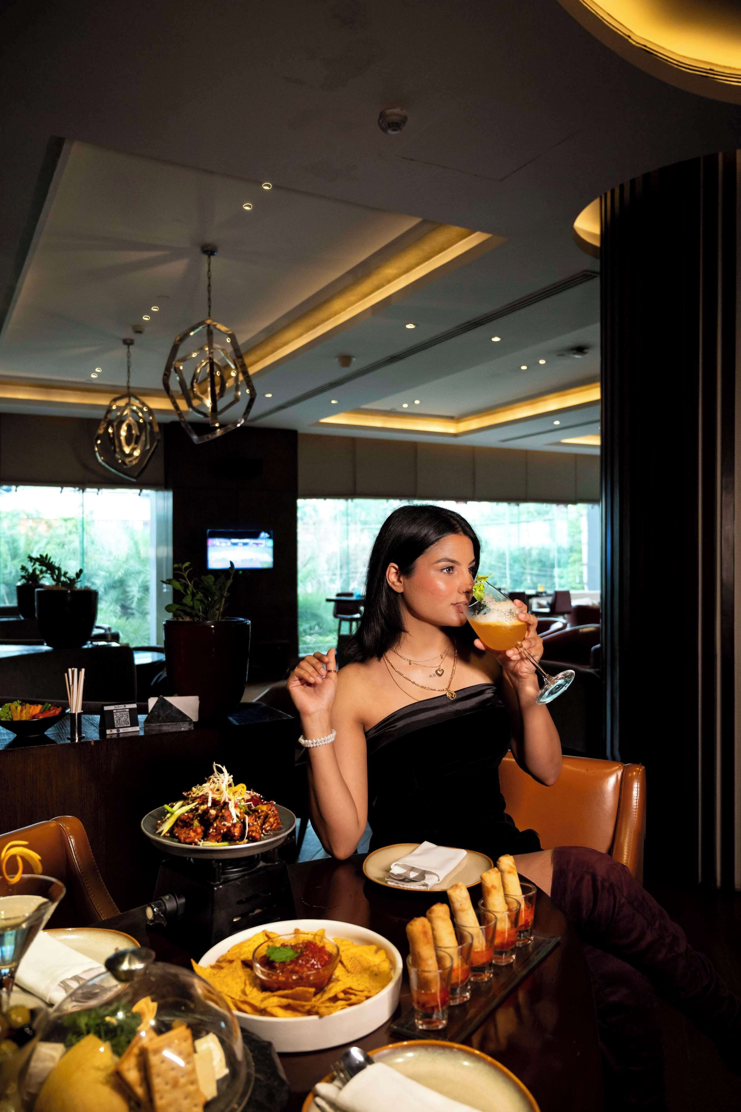 A woman seated at a table with a meal, including nachos and a drink, in a modern dining area with pendant lighting and a view of greenery outside.