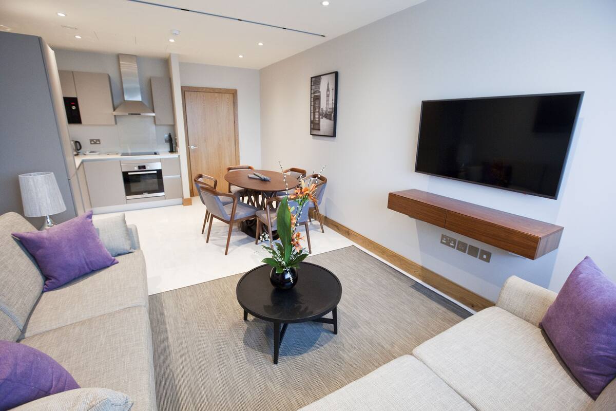 A modern living room with a sofa set, a coffee table with a vase, a dining area with chairs, a TV mounted on the wall, and a kitchen area with cabinets and a stove.