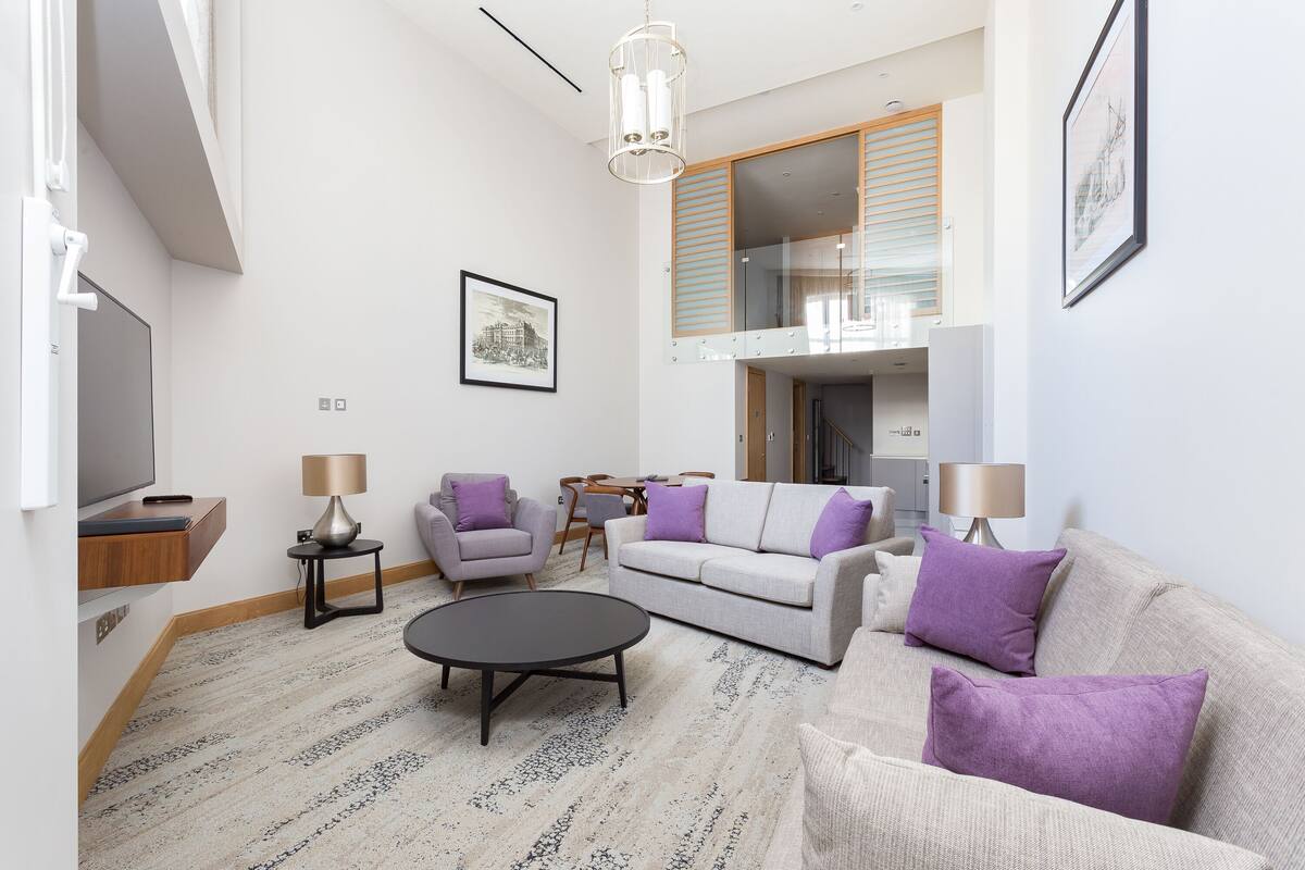 A modern living room with a sofa, armchairs, a coffee table, and a TV. There is a staircase leading to an upper level.