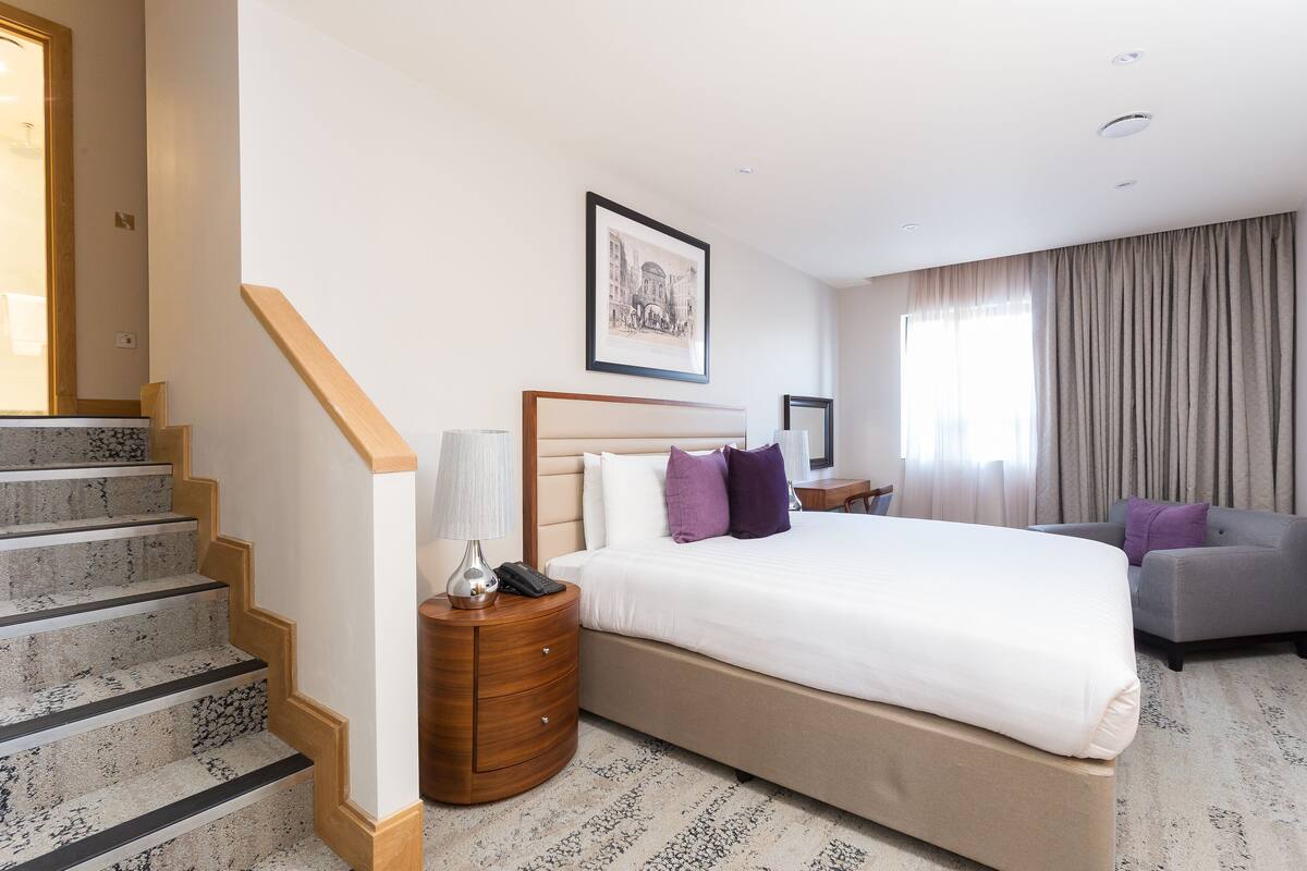 A hotel room with a large bed, a sofa, a nightstand, a lamp, a framed picture on the wall, and a staircase.