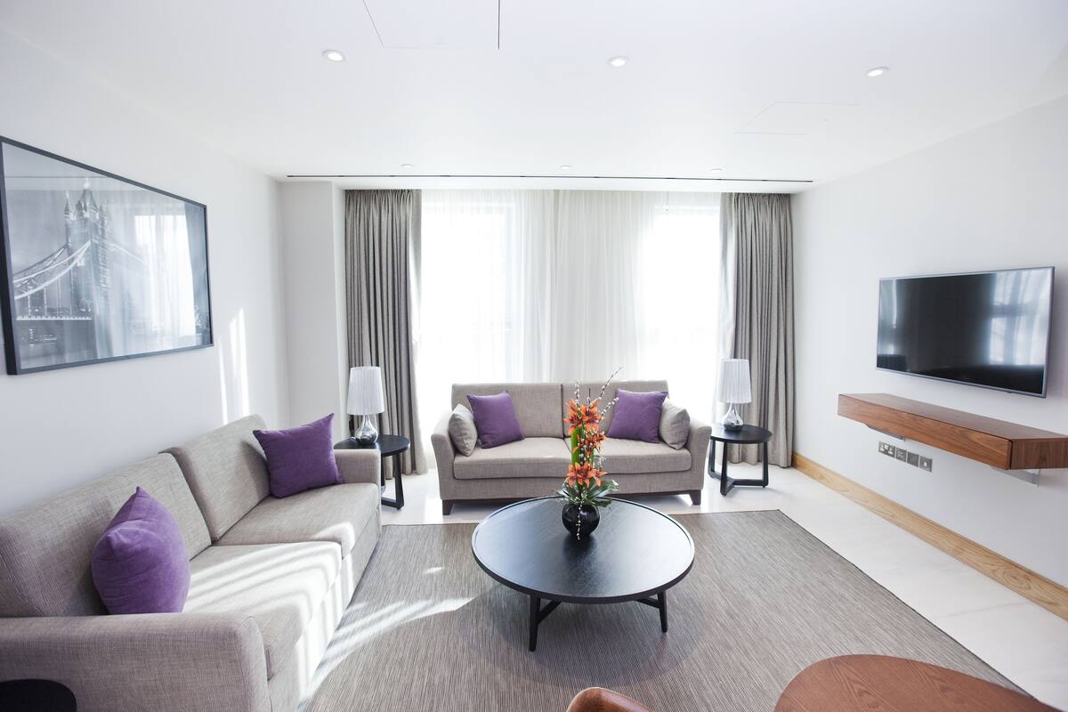 A modern hotel room with a sofa, a coffee table with a vase, a TV, and a large window with curtains.