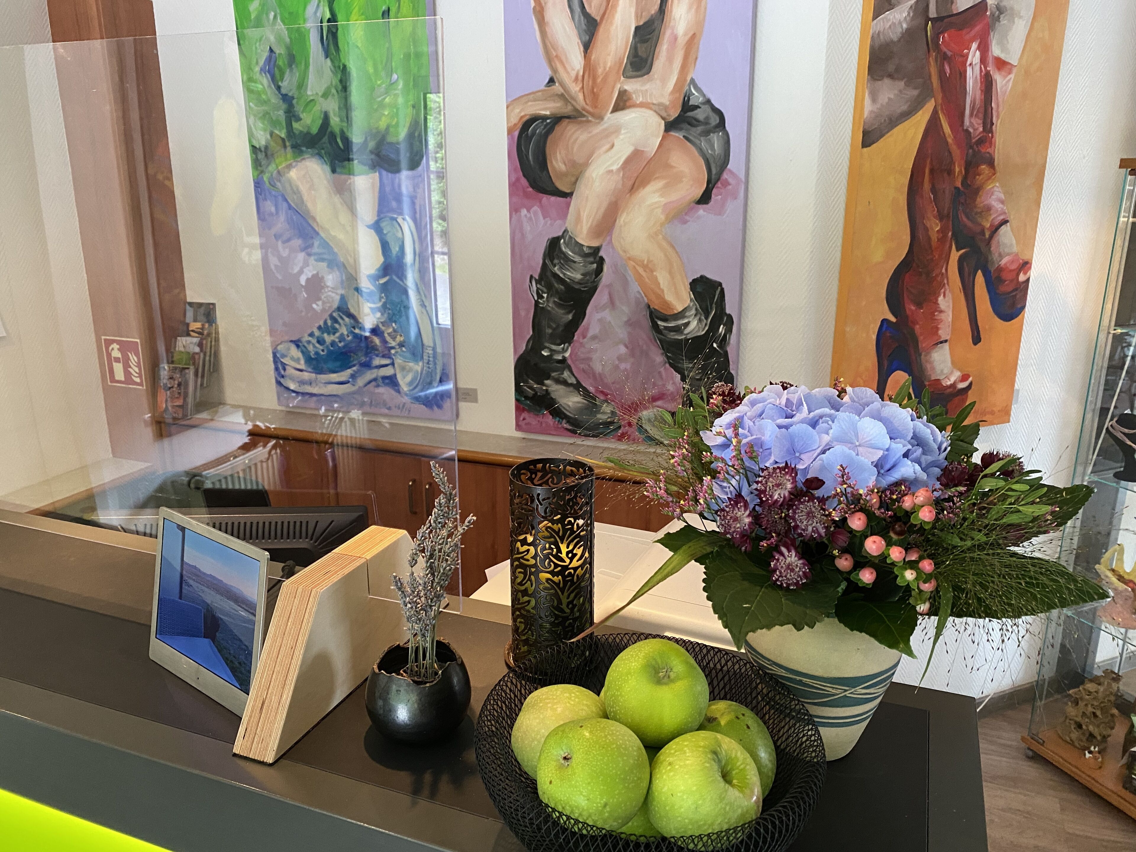 Un mostrador de recepción con una tableta, un florero con flores, un cuenco de manzanas y obras de arte en la pared.