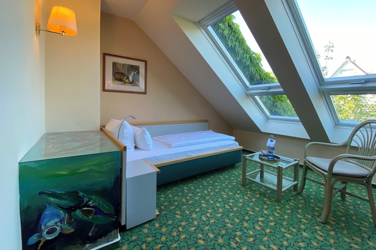Une pièce comprenant un lit, une petite table, une chaise, un tableau et une fenêtre donnant sur un paysage verdoyant.
