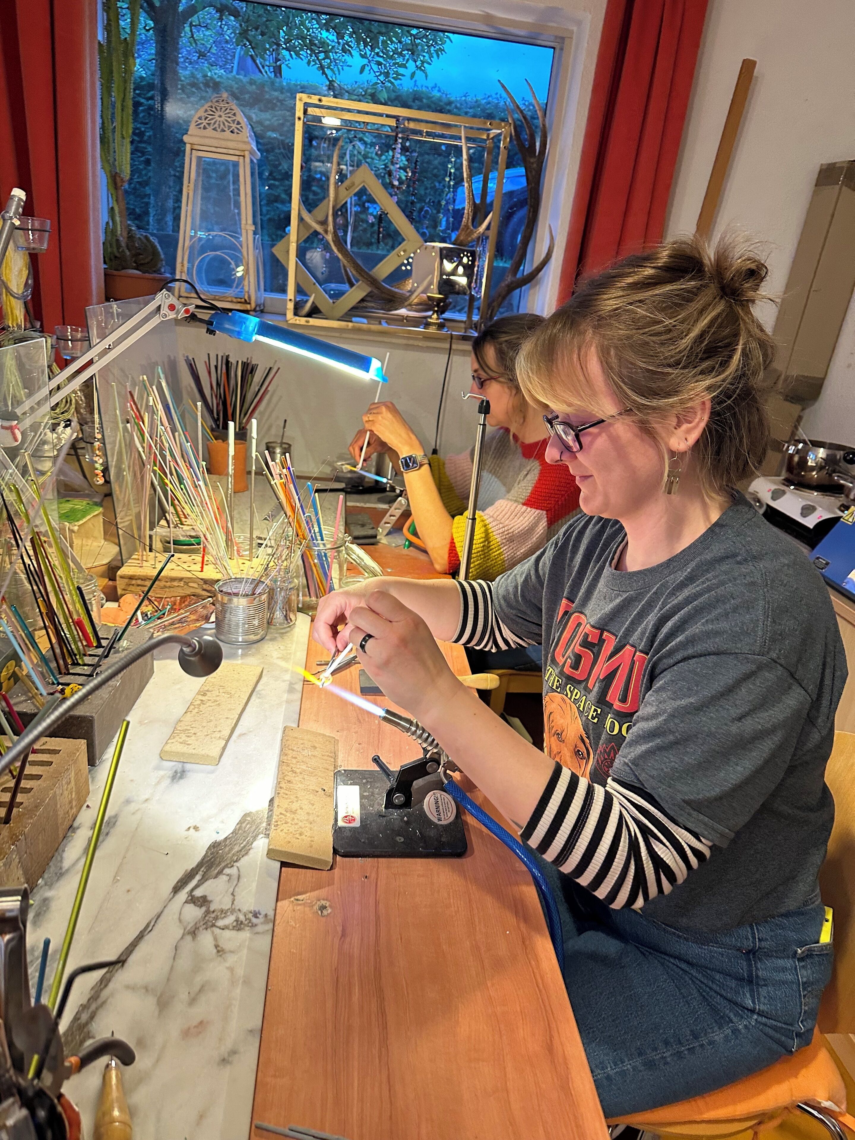 A person working at a desk with various tools and materials, a window with a view of trees, and a red curtain.