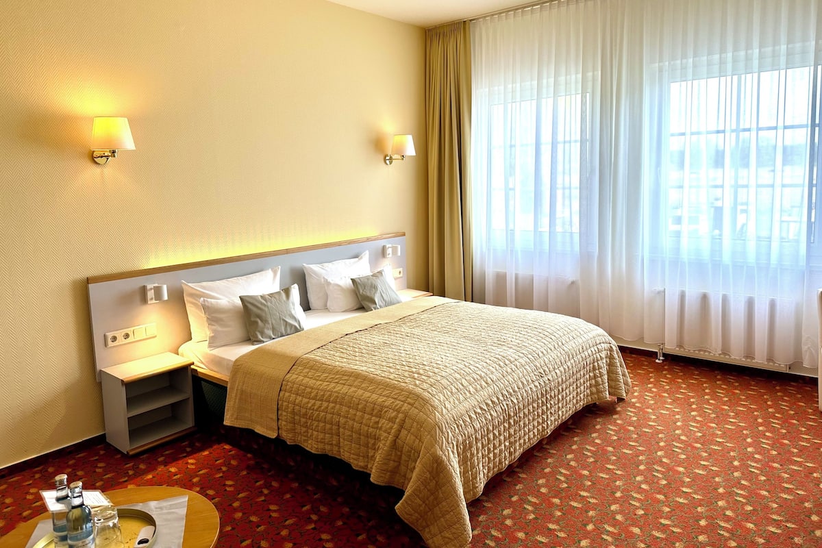 Une chambre d’hôtel avec un grand lit, deux tables de chevet, une petite table d’appoint, une table ronde avec un plateau, et une fenêtre avec des rideaux légers.