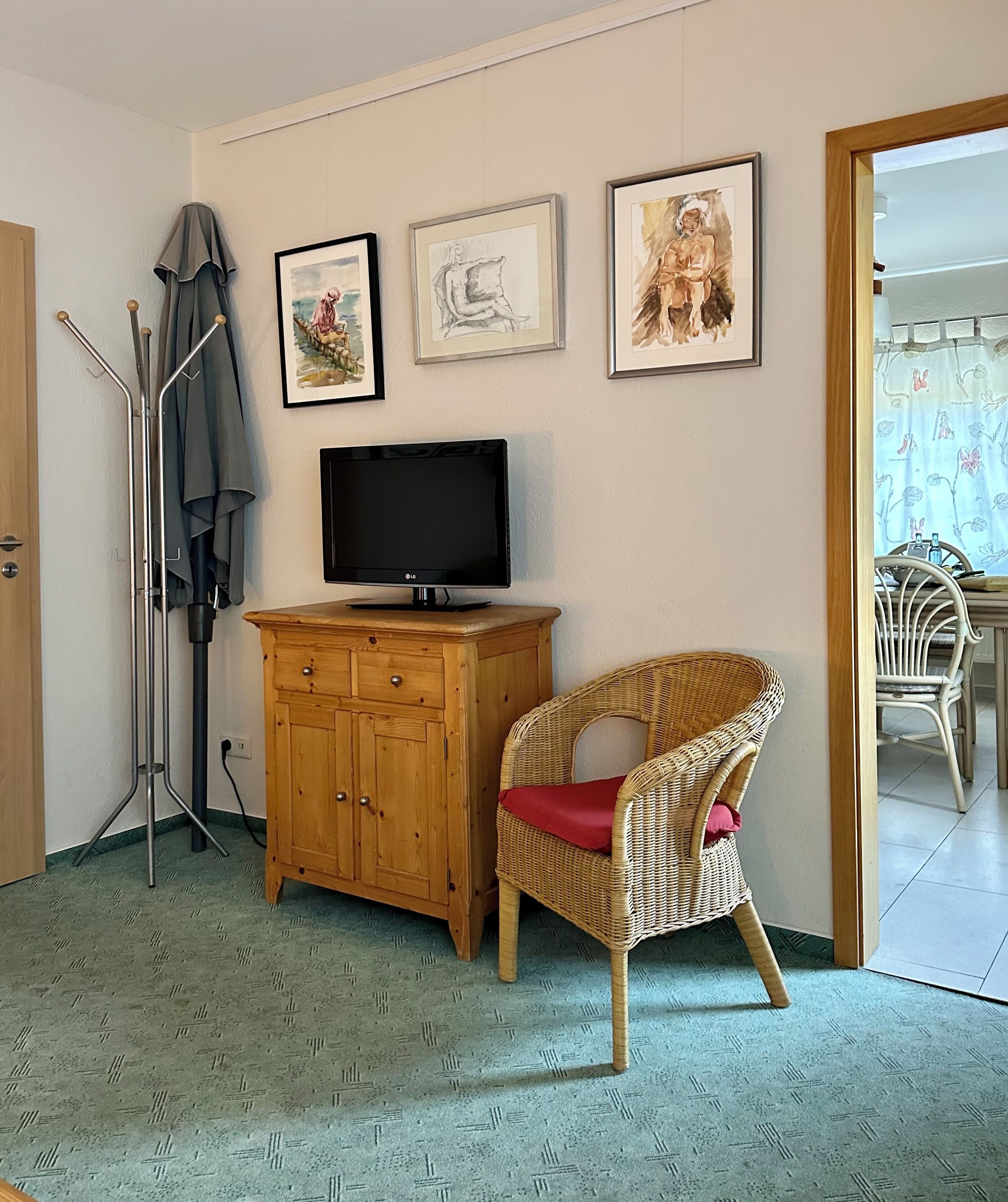 Ein Zimmer mit einem Holzkasten, einem Korbstuhl mit rotem Kissen, einem Fernseher und drei gerahmten Kunstwerken an der Wand.