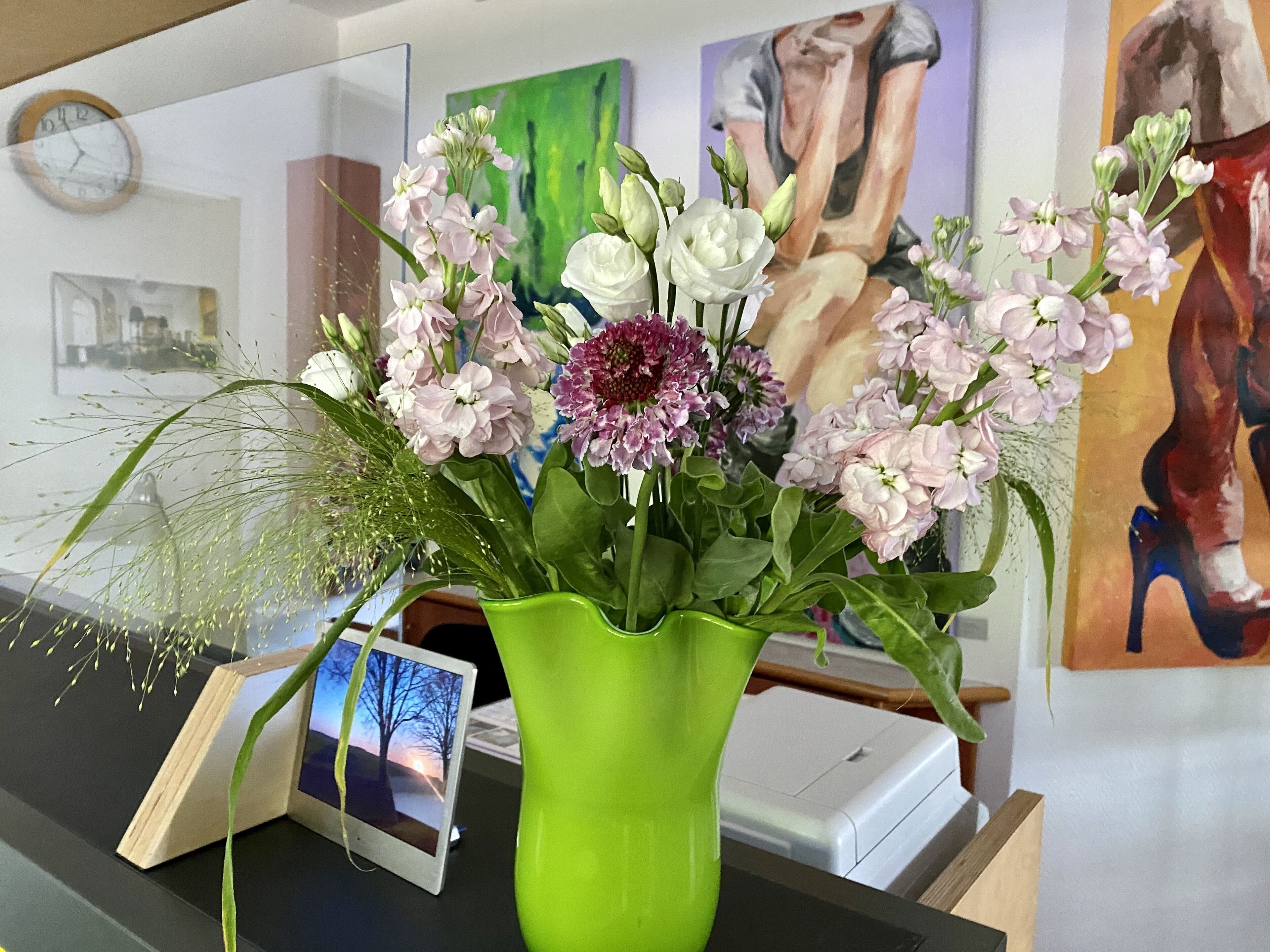 Un bureau sur lequel se trouve un vase vert contenant des fleurs, une horloge et une photo encadrée.