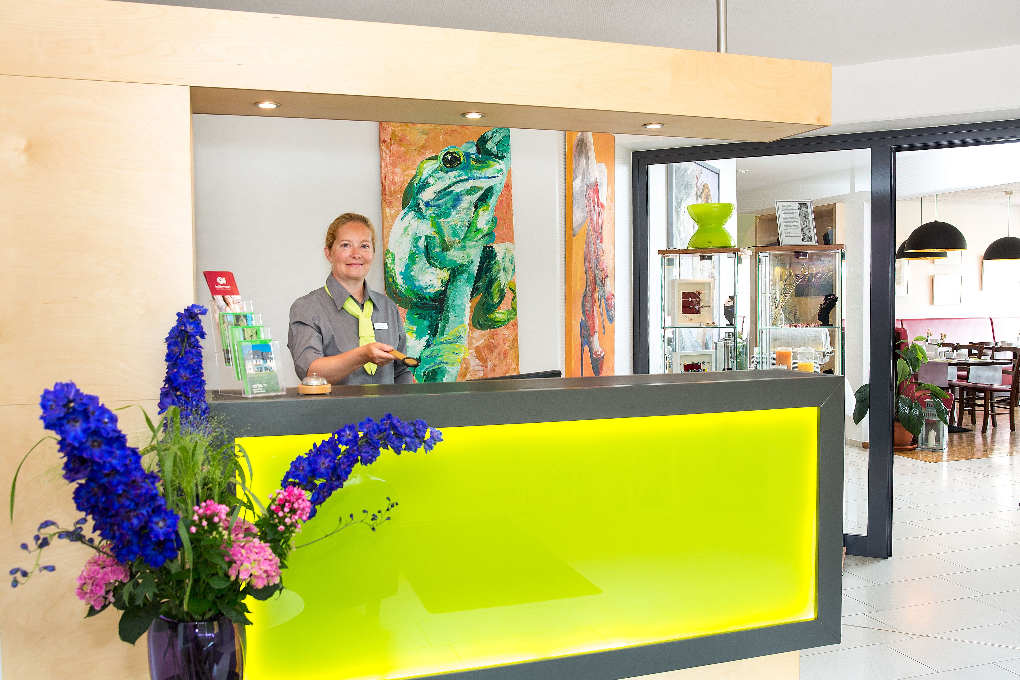 Een receptiebalie met een receptioniste, een kleurrijk schilderij van een kikker en een vaas met bloemen.