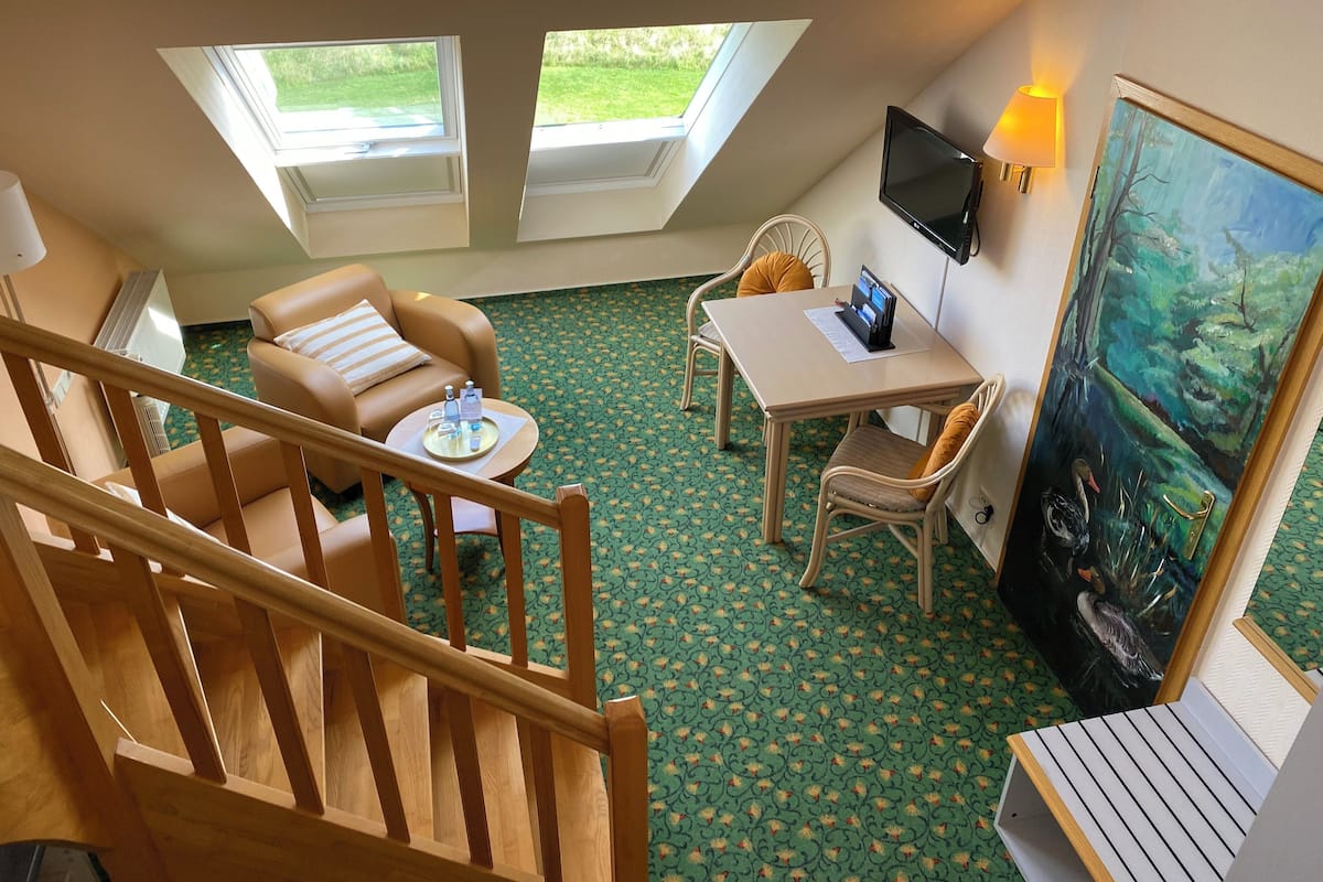 Une pièce avec un plafond haut, un escalier en bois, un canapé, une table avec un ordinateur portable, un tableau et deux fenêtres donnant sur un paysage verdoyant.