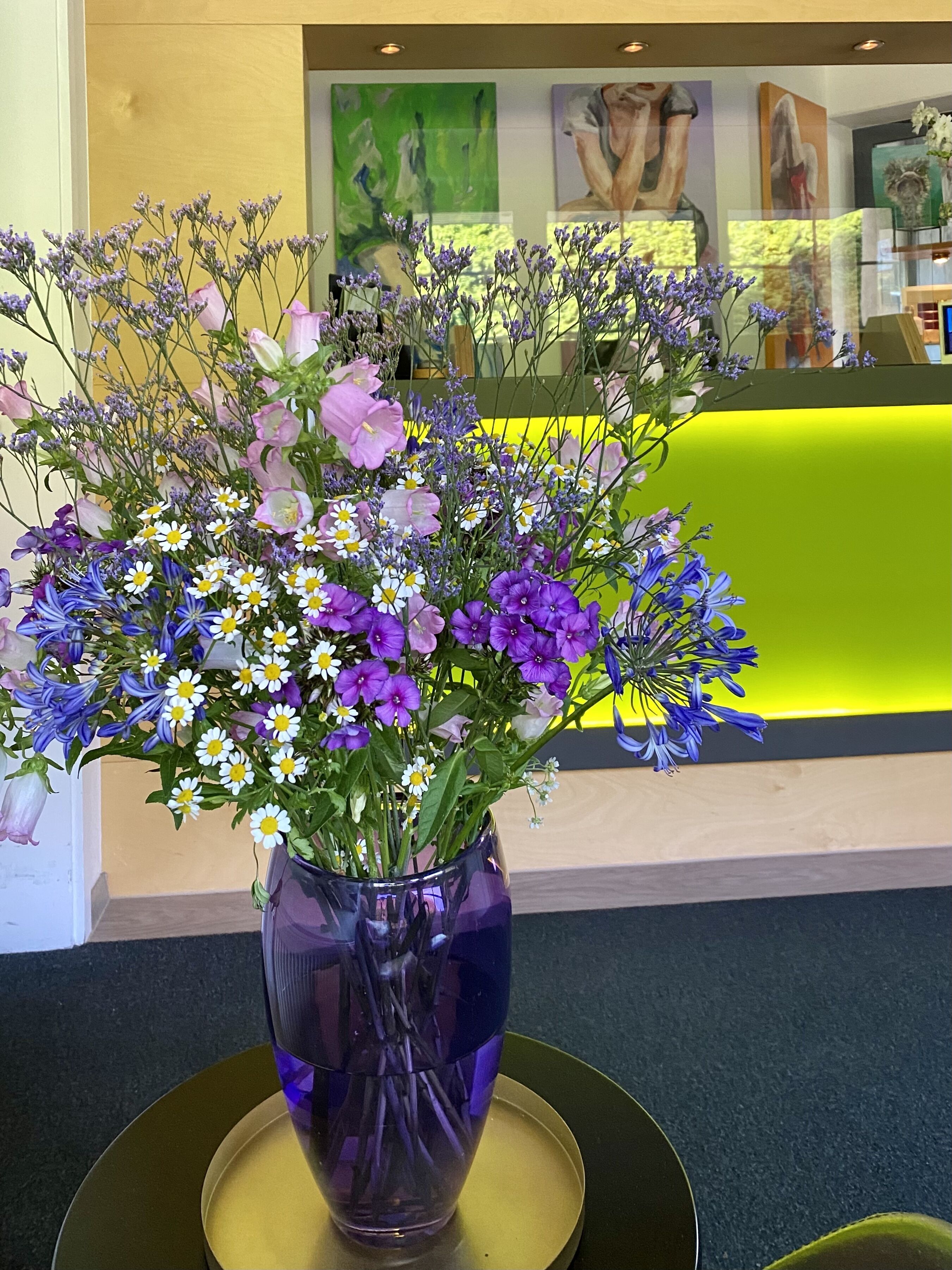 Eine Vase mit verschiedenen Blumen auf einem Tisch in einem Raum mit gelber Wand und Kunstwerken.