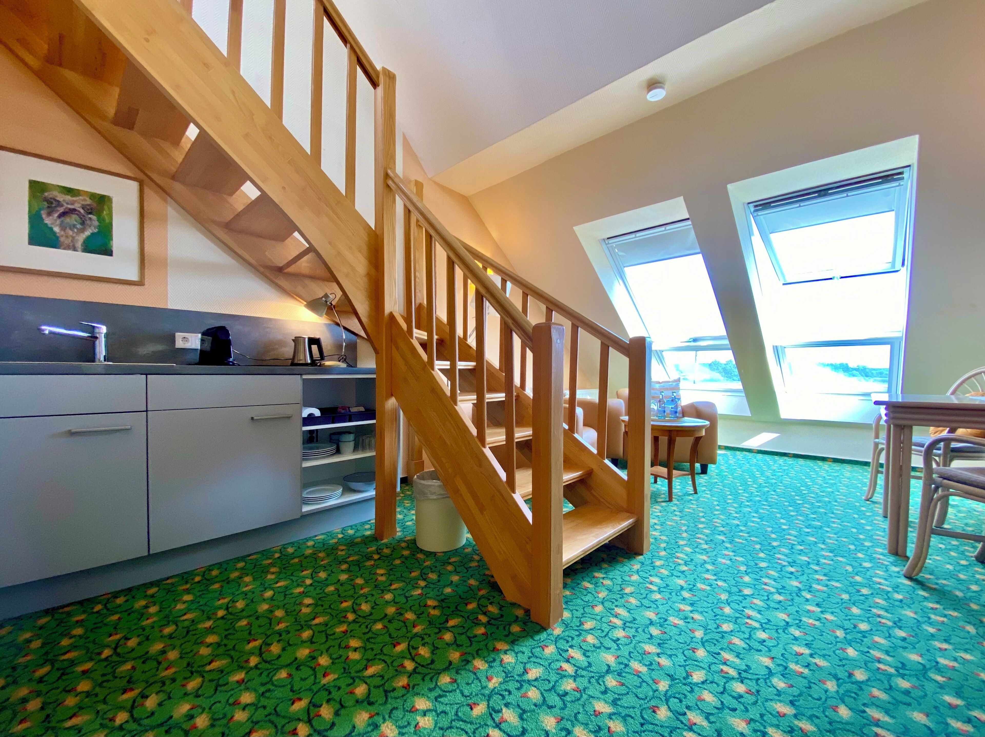 A modern kitchen with a wooden staircase, a dining area with a table and chairs, and a large window providing natural light.