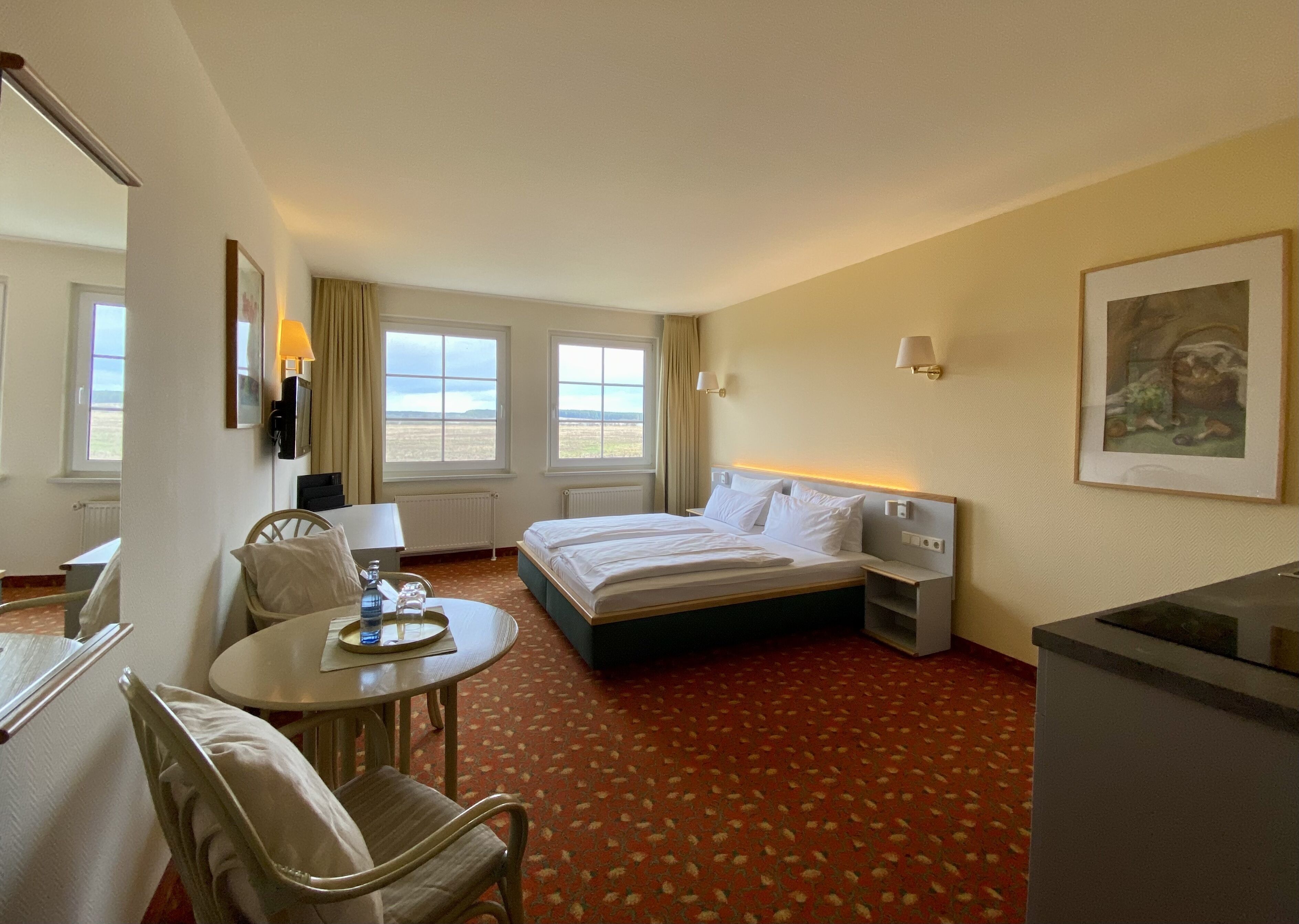 A hotel room with a bed, a desk, a chair, a table with a tray, and a painting on the wall.
