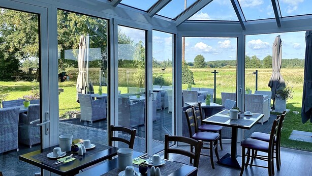 A conservatory with glass walls, outdoor seating, and a view of a grassy field. - Hotel am Müritz-Nationalpark (Waren (Müritz))