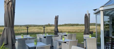 Eine Terrasse mit Korbmöbeln, Tisch und Sonnenschirm, mit Blick auf eine Wiese.