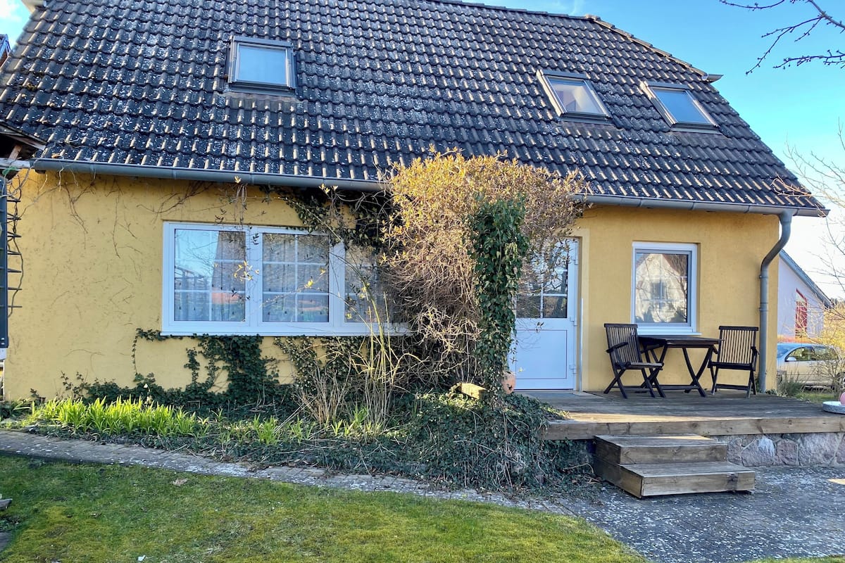 Une maison jaune avec un toit en tuiles, deux fenêtres, une porte, un petit porche avec une table et des chaises, et une cour couverte d’herbe.