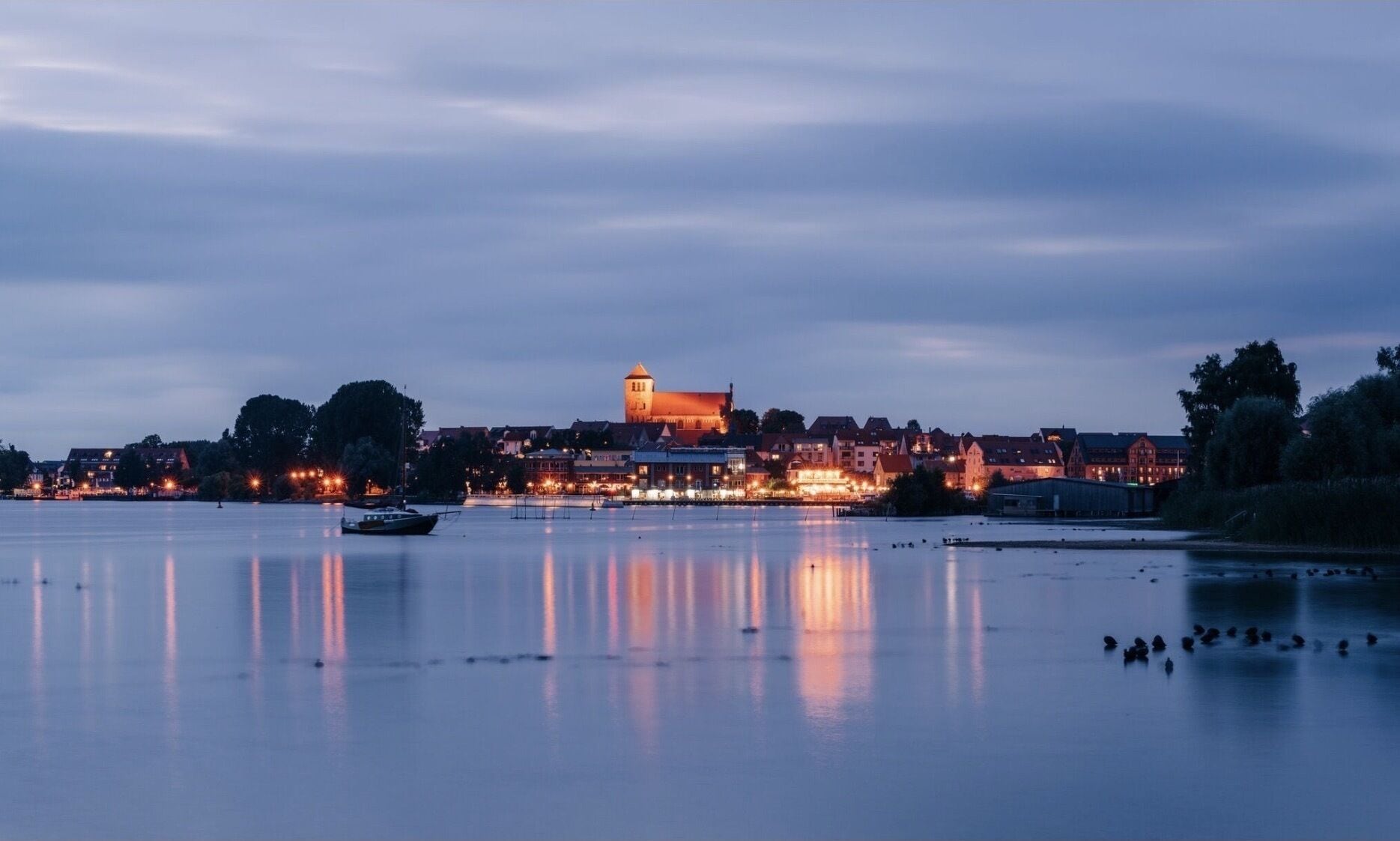 Une rivière paisible, avec une ville sur la rive opposée, caractérisée par une tour imposante et des bâtiments illuminés.