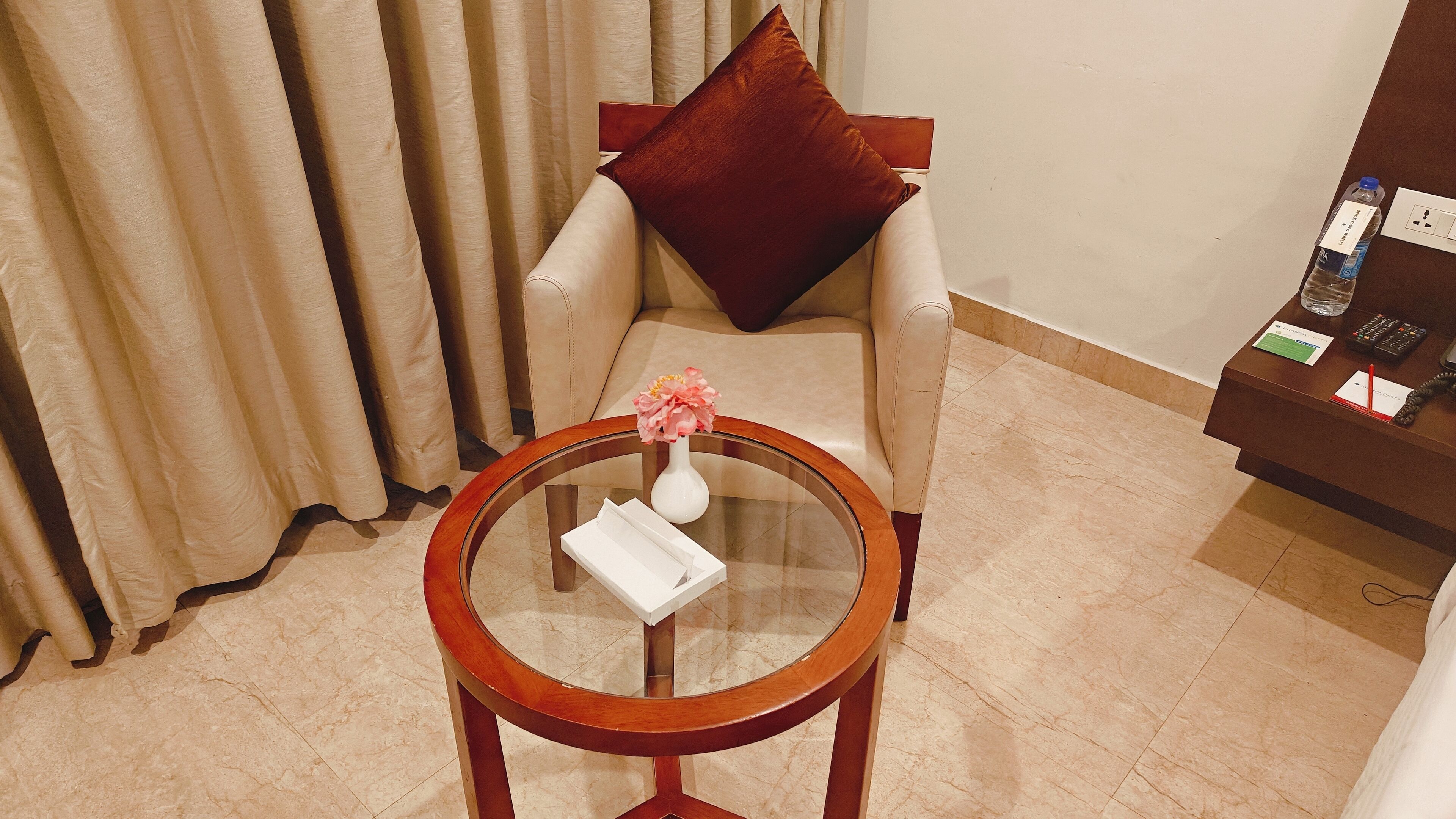 Habitación de hotel con una mesa redonda de madera, un jarrón blanco con flores, un cojín marrón sobre un sillón color beige y una mesita auxiliar marrón con una botella y un control remoto.