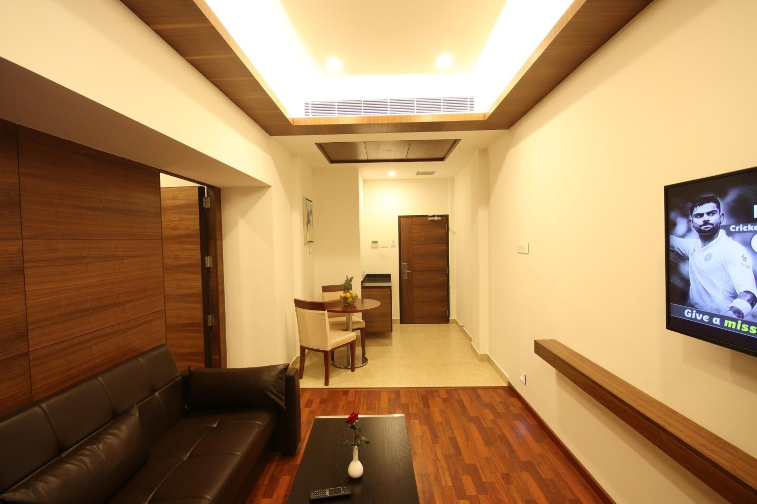 Un salón moderno con suelo de madera, un sofá de cuero negro, una mesa de centro con un jarrón y un televisor en la pared mostrando a un jugador de criquet.