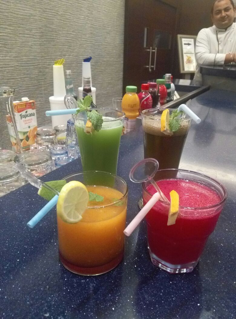 Uma área de bar com diversas bebidas, incluindo um líquido verde, um laranja e um vermelho, e uma pessoa ao fundo.