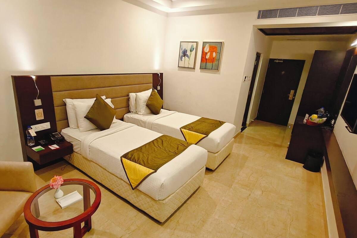 A hotel room with two beds, a nightstand, a small table with a vase, and a chair.