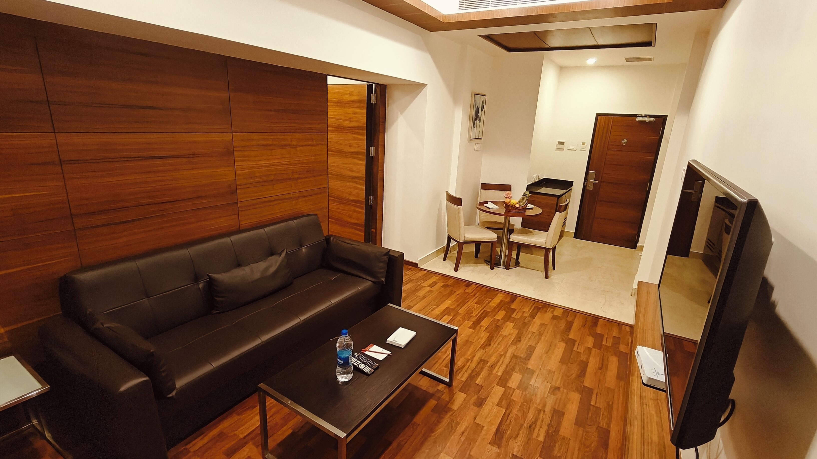 A modern living room with a black leather sofa, a wooden wall panel, a dining area with a table and chairs, and a flat-screen TV.