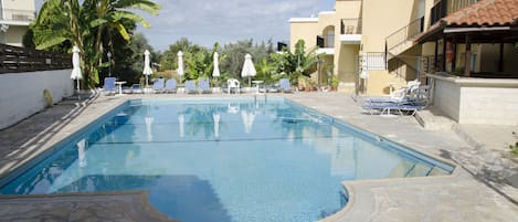 A swimming pool with lounge chairs and umbrellas, surrounded by palm trees and buildings.