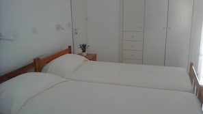 Ein Schlafzimmer mit zwei Einzelbetten, einem Holz-Kopfteil, einem Nachttisch mit Pflanze und einem Kleiderschrank mit mehreren Fächern.