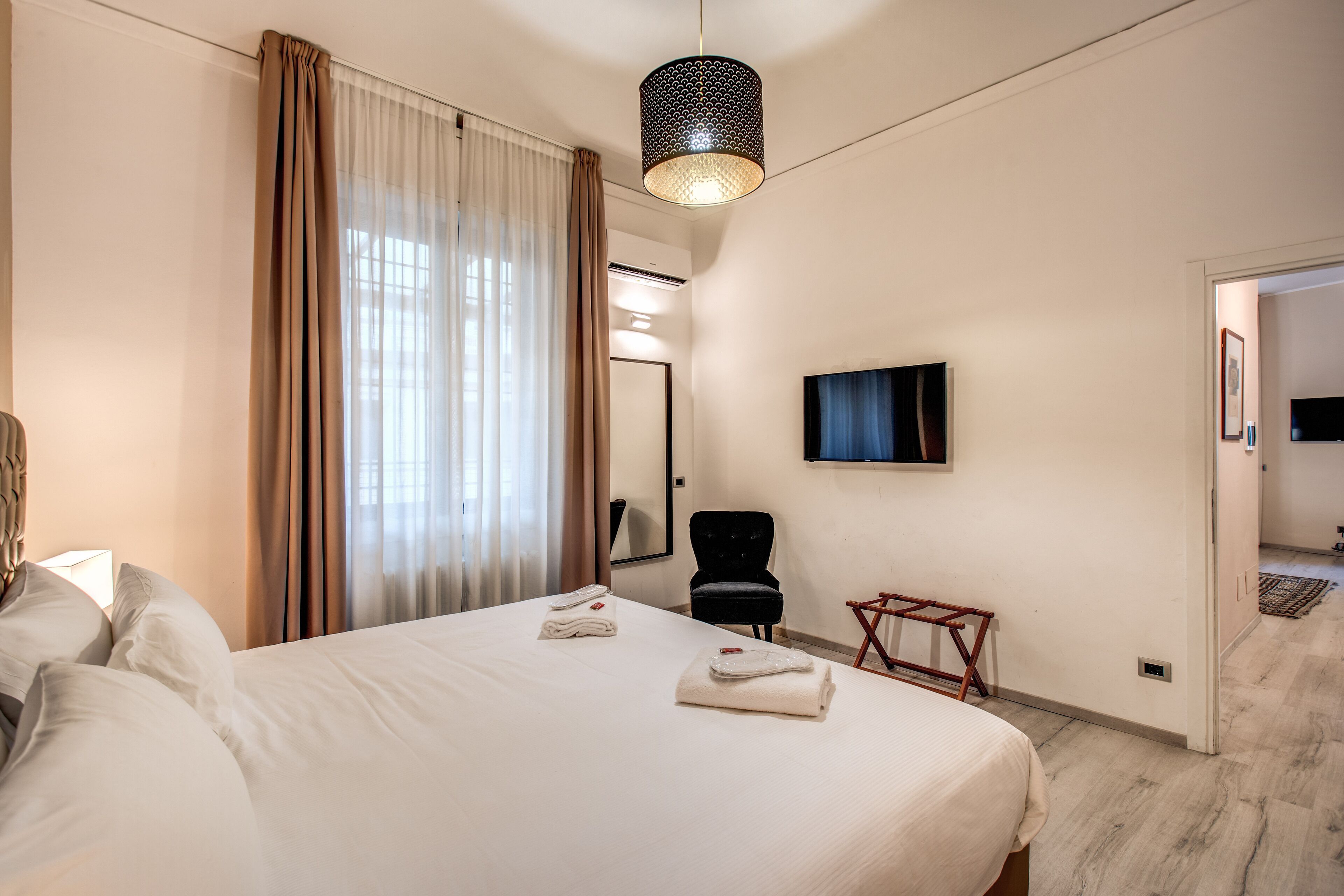Ein Hotelzimmer mit Bett, Sessel, Wandfernseher und einer Tür zu einem weiteren Raum.