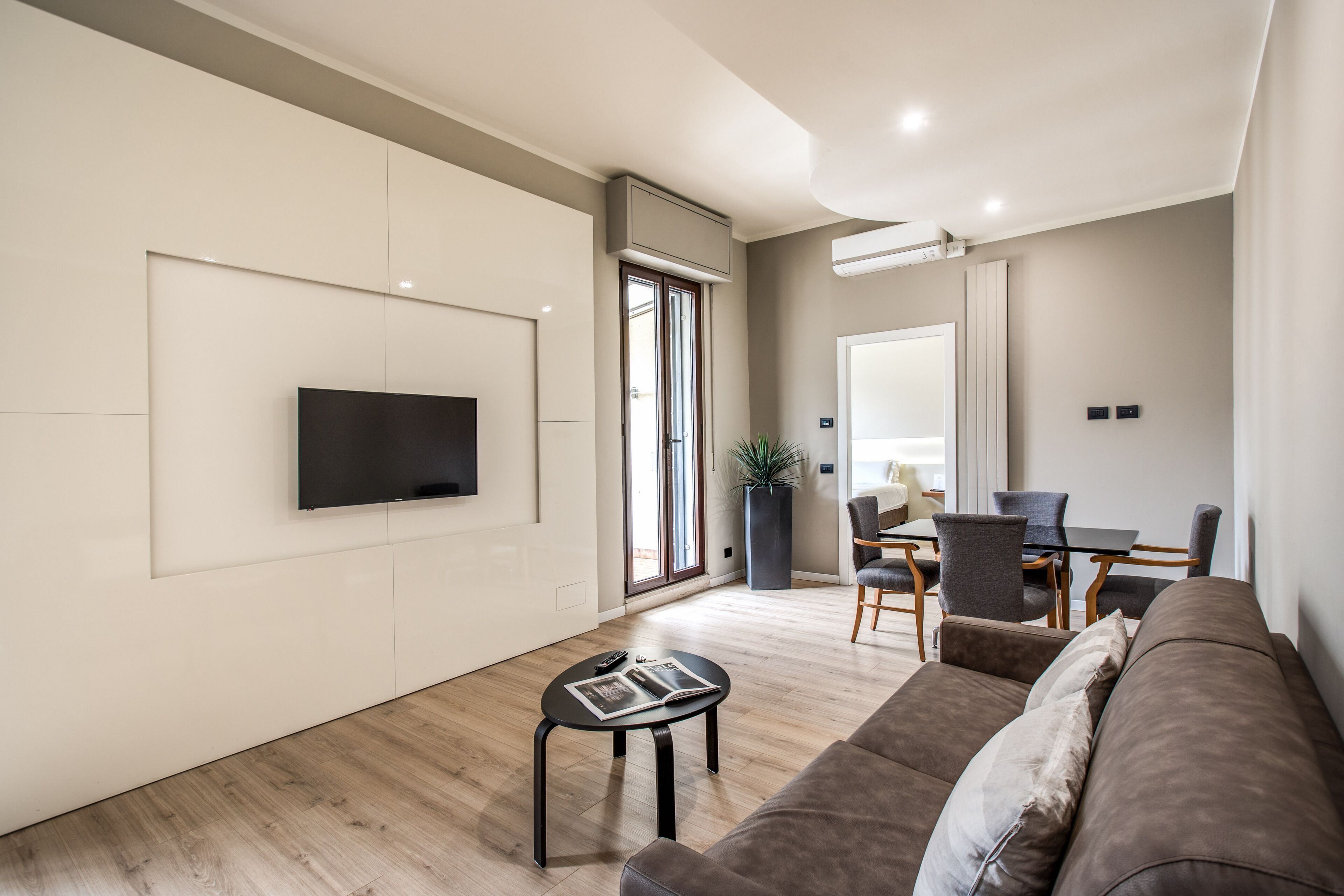 A modern living room with a flat-screen TV mounted on the wall, a brown sofa, a small coffee table with a magazine, and a dining area with a table and chairs.